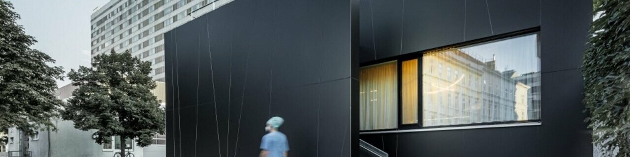 Frontalansicht der zentralen Notaufnahme der Klinik Landstraße in der Juchsgasse in Wien wurde mit PREFABOND Aluminium Verbundplatten in der Farbe schwarzgrau verkleidet. Im Bild ist eine Krankenschwester zu sehen.