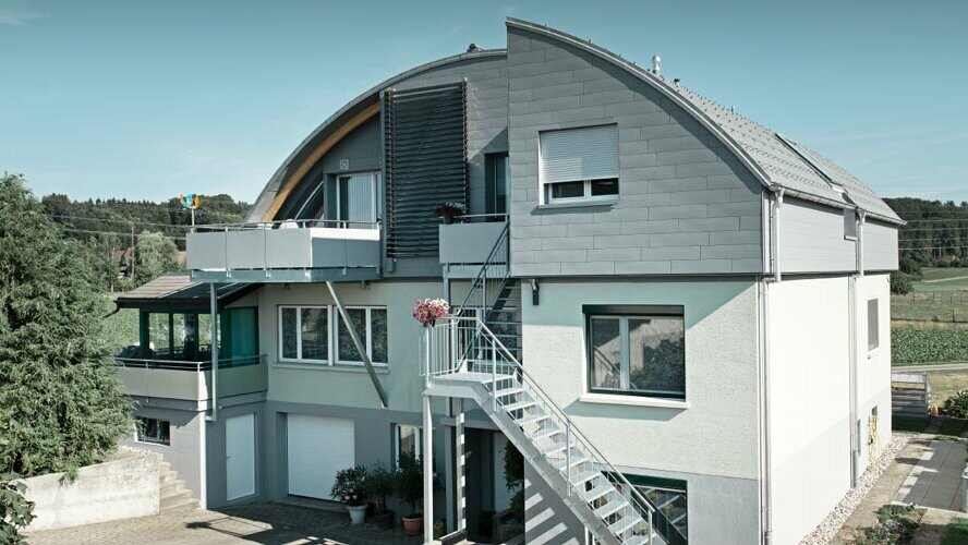 Obytný dům s valenou střechou pokrytou PREFA falcovanými střešními taškami a svitkovým plechem Prefalz světle šedé barvy P.10.