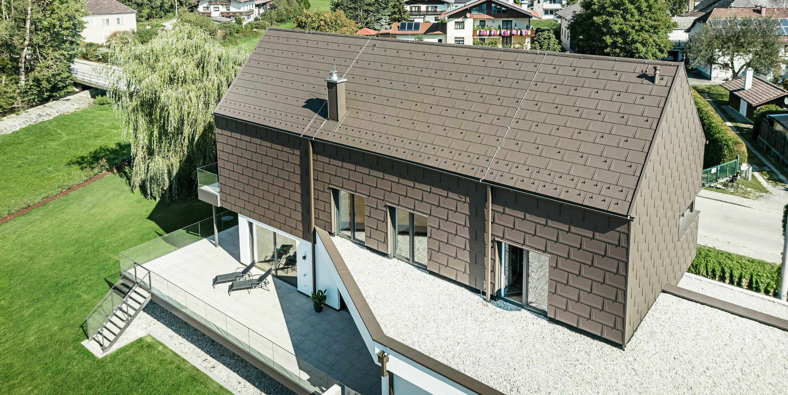 Pohled z ptačí perspektivy na unikátní rodinný dům v rakouském Neukirchenu opláštěný střešními panely PREFA R. 16 v barvě P.10 hnědá. Maloformátové hliníkové prvky zdobí sedlovou střechu a horní patro moderního domu. Odvodnění střechy z PREFA ve stejné barvě je provedeno přes čtvercovou trubku i boxový okap a sběrný box na vodu. Velká okna umožňují pohled zevnitř budovy na klidnou rezidenční krajinu Neukirchenu s četnými zelenými korunami stromů.