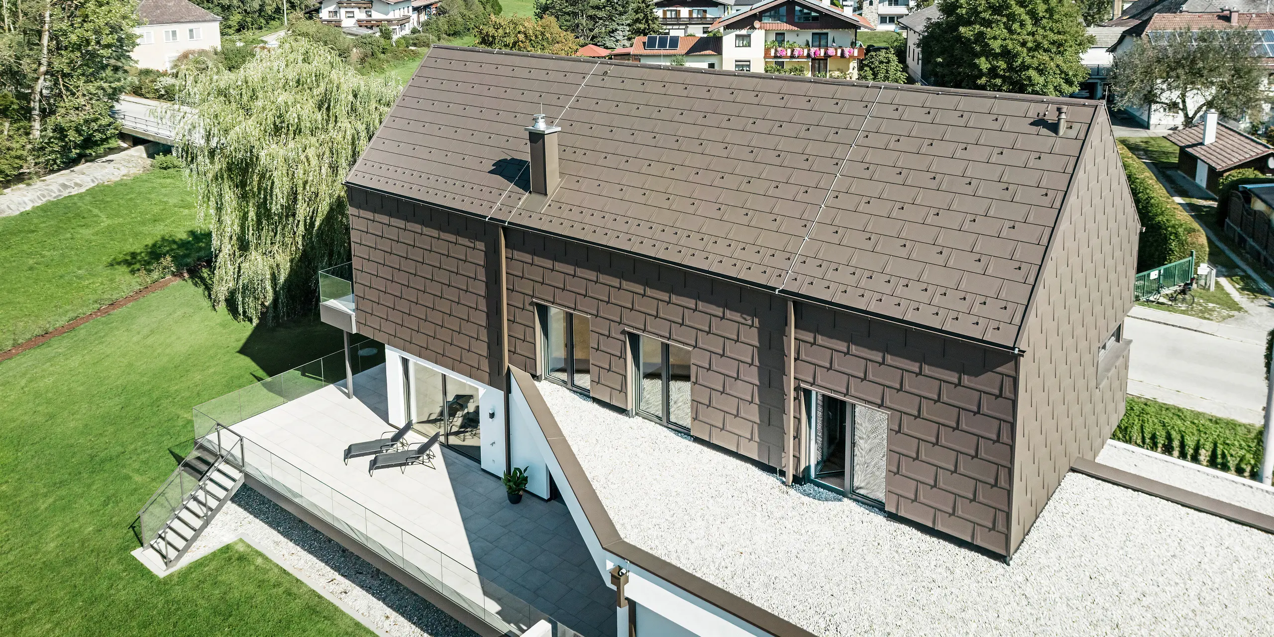 Pohled z ptačí perspektivy na unikátní rodinný dům v rakouském Neukirchenu opláštěný střešními panely PREFA R. 16 v barvě P.10 hnědá. Maloformátové hliníkové prvky zdobí sedlovou střechu a horní patro moderního domu. Odvodnění střechy z PREFA ve stejné barvě je provedeno přes čtvercovou trubku i boxový okap a sběrný box na vodu. Velká okna umožňují pohled zevnitř budovy na klidnou rezidenční krajinu Neukirchenu s četnými zelenými korunami stromů.