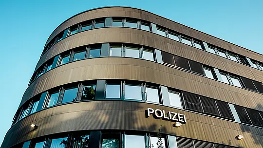 Dramatický pohled zdola na zakřivenou novostavbu policejní stanice 43 v Hamburku: Výrazná hliníková fasáda PREFA z eloxovaných extrudovaných profilů „Zacke“ ve střední bronzové barvě je architektonickým poutačem. Jemná, rytmická struktura vytváří dynamické světelné a stínové vzory a zdůrazňuje plynulé křivky budovy. Hliník PREFA nabízí vysokou odolnost proti povětrnostním vlivům, ochranu proti korozi a dlouhou životnost – udržitelné a nenáročné řešení pro precizní a moderní fasádní architekturu.