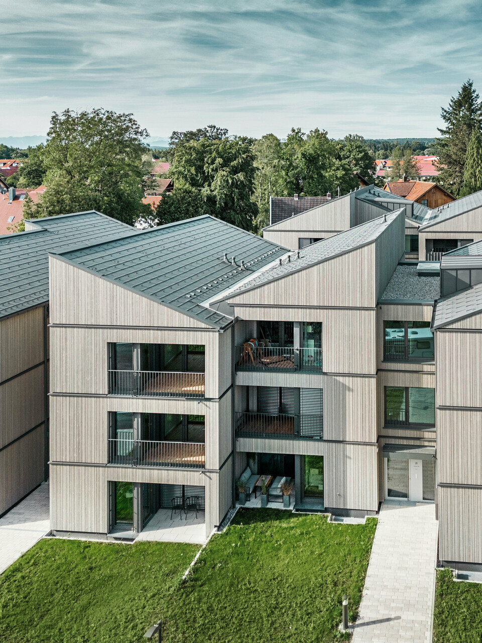 [Translate to Český:] Luftaufnahme des modernen Mehrfamilienhauses am Schmucker-Areal in Utting am Ammersee, mit nachhaltiger Holzhybridbauweise und eleganten Holzfassaden. Die Gebäude sind mit langlebigen PREFA Dachplatten R.16 in P.10 Dunkelgrau ausgestattet. Diese innovative Architektur, geplant von WWA Architekten aus München, zeichnet sich durch klare Linien und harmonische Integration in die grüne Umgebung aus. Die großzügigen Balkone und großen Fenster sorgen für eine helle und freundliche Wohnatmosphäre, während die hochwertigen Materialien für Langlebigkeit und Wetterbeständigkeit sorgen.