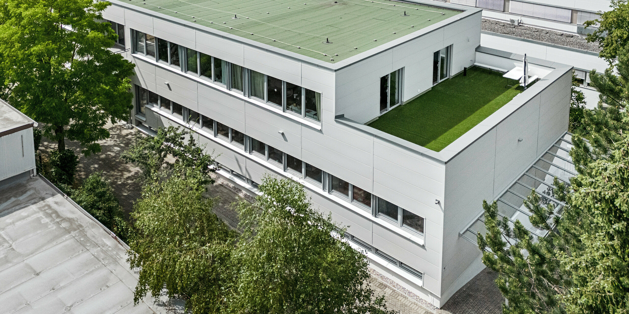Pohled z ptačí perspektivy na budovy se zelenou střechou a terasou. Budova je obložená velkoformátovými fasádními lamelami Siding 500 a 600 od firmy PREFA. 