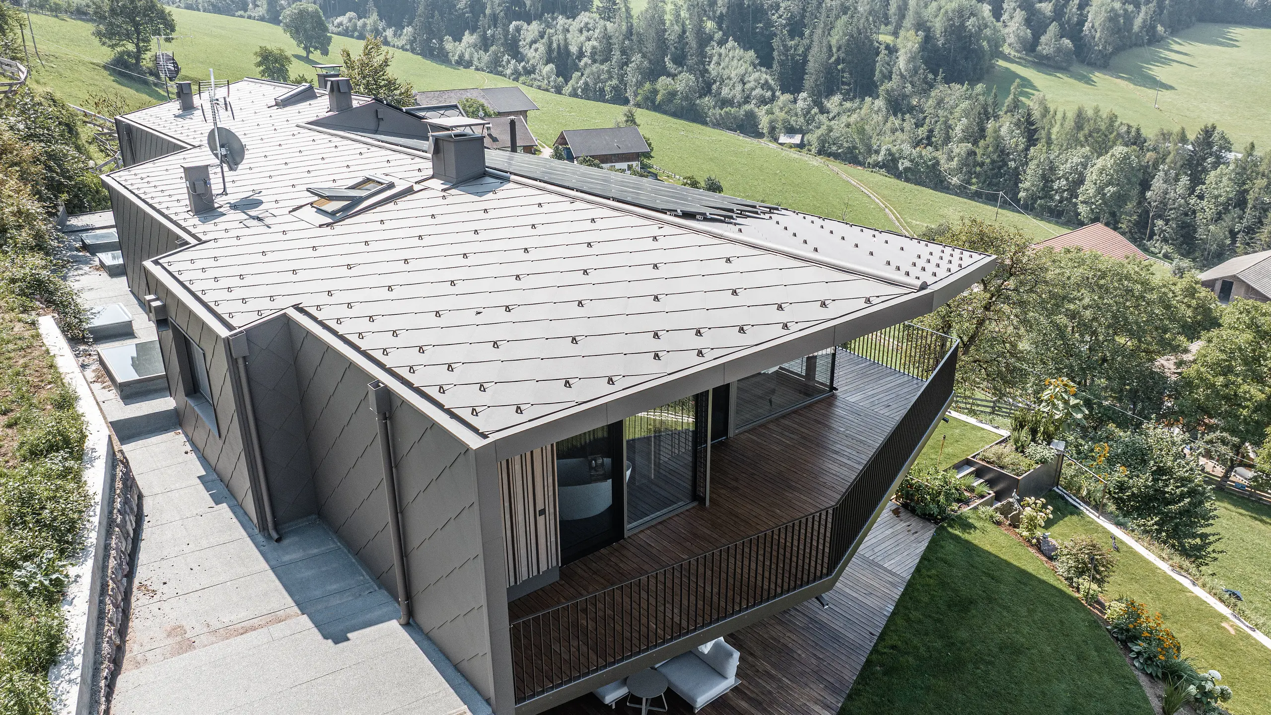 Pohled na zmodernizovaný dvojdům v Möltenu v Jižním Tyrolsku vybavený odolnými falcovanými a falcovanými šablonami 44x44 v barvě P.10 tmavě hnědé. Design střechy a fasády kombinuje robustní hliník s dřevěnými prvky, obklopený malebnou krajinou s rozsáhlými výhledy na zalesněné kopce. Redesign plynule integruje zrekonstruovanou stávající budovu a nově vybudovaný areál do harmonického celku se zaměřením na dřevo jako stavební materiál.