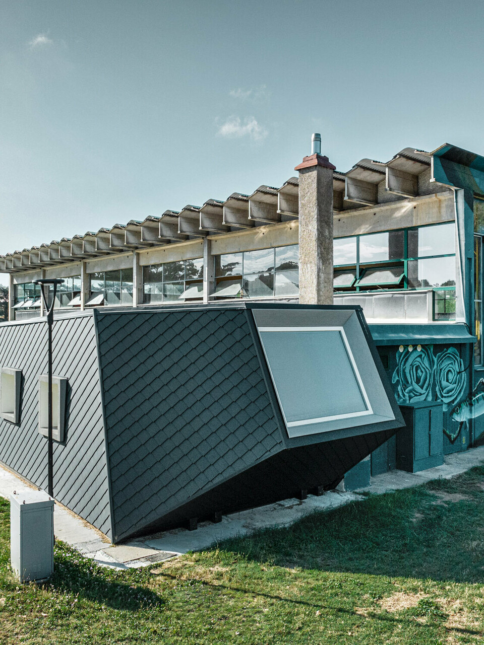 Die Turnhalle Rodriguez und ihr neuer Zubau mit der Rautenschuppenhaut aus PREFA Aluminium von der Seite; der Rasen erstreckt sich davor und zur Seite, im Hintergrund ist ein Teil eines Sportplatzes zu sehen.