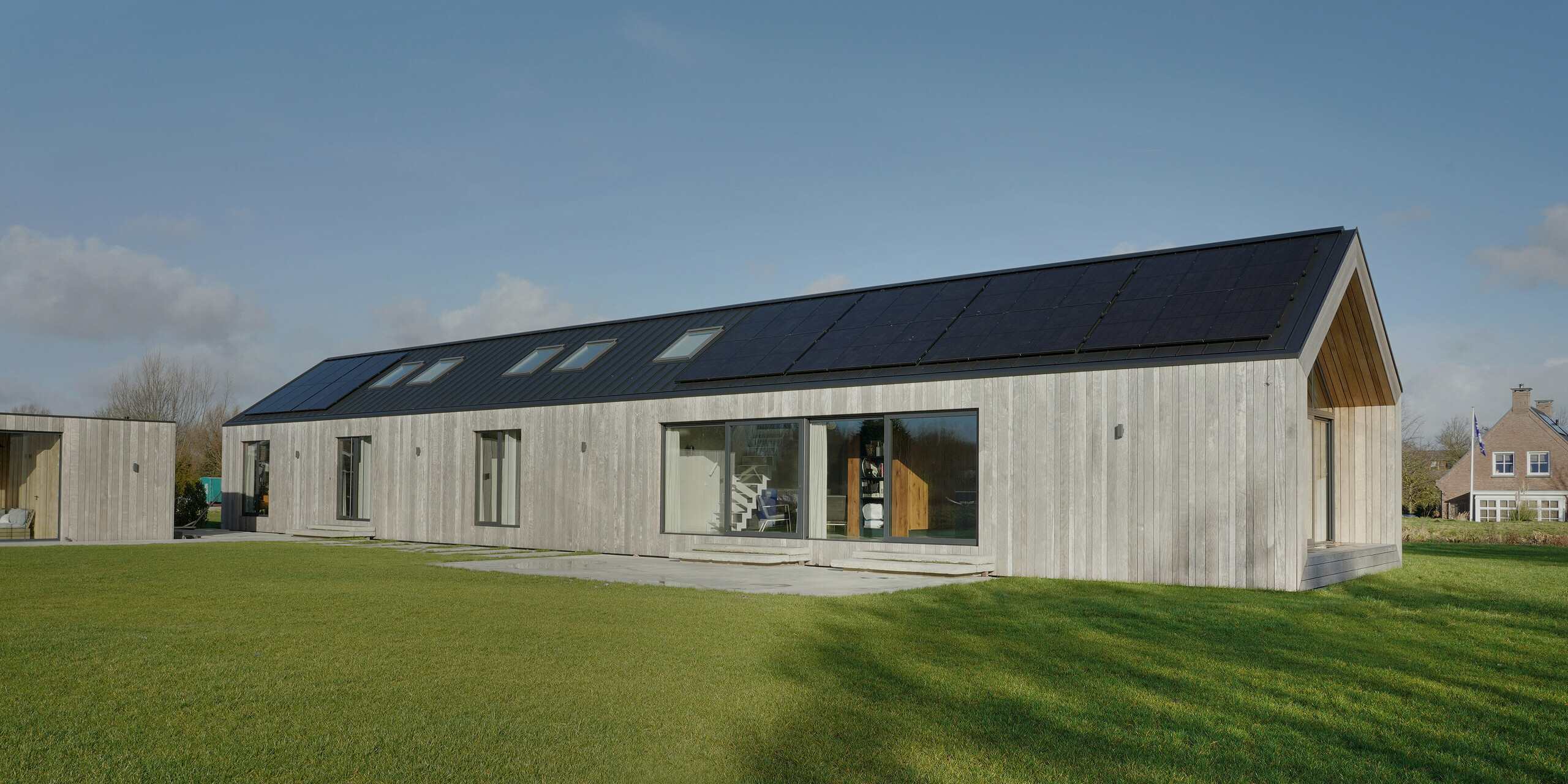 Weitwinkelaufnahme eines modernen Bungalows in Uithoorn, Niederlande, mit PREFALZ Winkelstehfalz-Eindeckung in Schwarzgrau und Photovoltaik-Anlage als Aufdach-Lösung. Das Bild zeigt das geräumige Gebäude mit großen Glasfronten und Holzverkleidung, umgeben von einem gepflegten Rasen. Die nachhaltige Bauweise kombiniert ästhetisches Design mit langlebigen, wetterfesten Materialien, ideal für umweltbewusste Bauvorhaben.