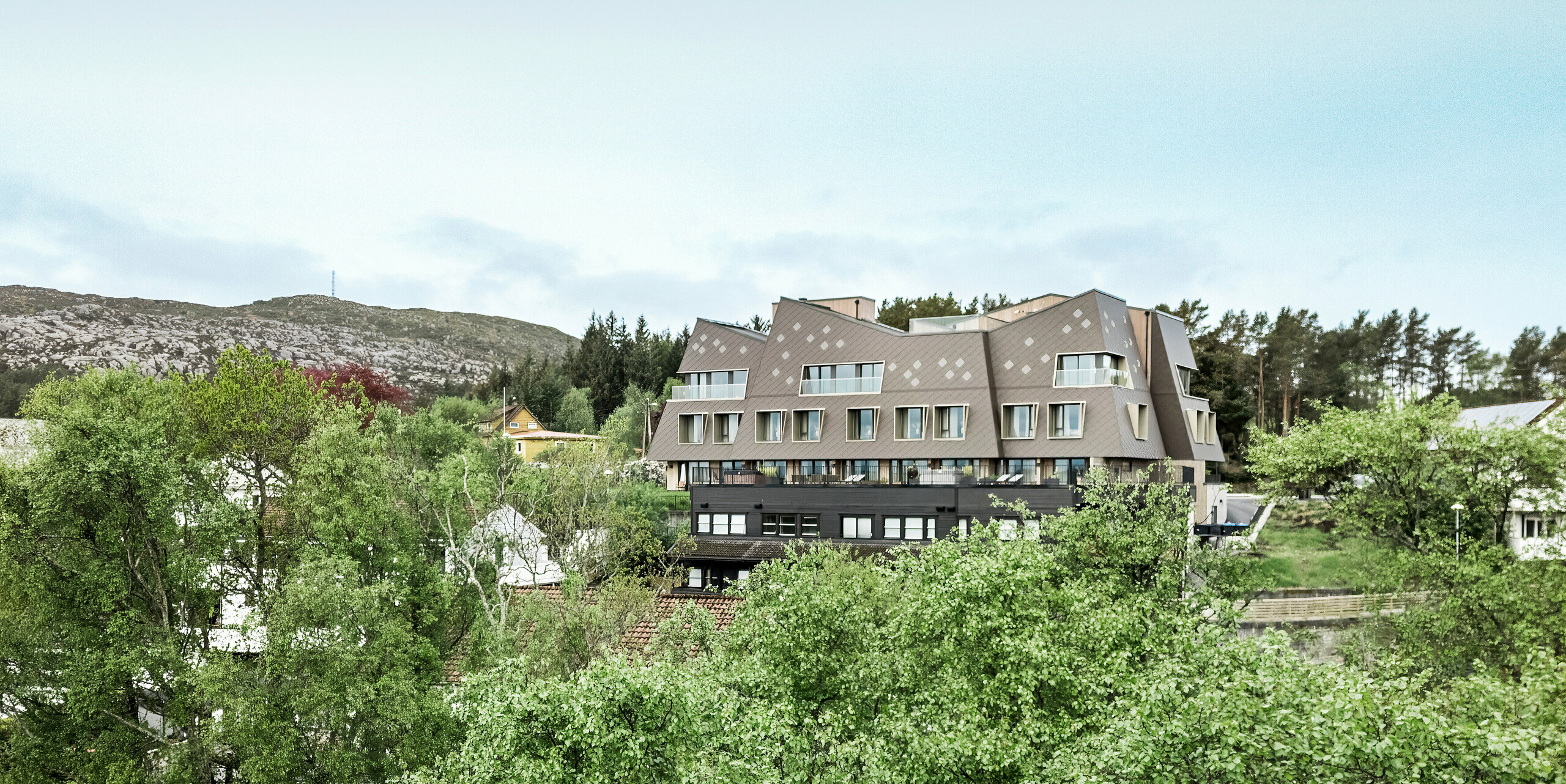 Vzdálený pohled na hotel Beckerwyc House v Bekkjarviku v Norsku zasazený do zelené kopcovité krajiny. Moderně vyhlížející budova je obložena fasádními šablonami PREFA 44 × 44 v barvě P.10  oříškové, které harmonicky zapadají do skalnatého okolí. Mimořádná architektura hotelu vyčnívá z okolních tradičních domů a přesto díky svému zemitému barevnému provedení dobře splývá s přírodou.