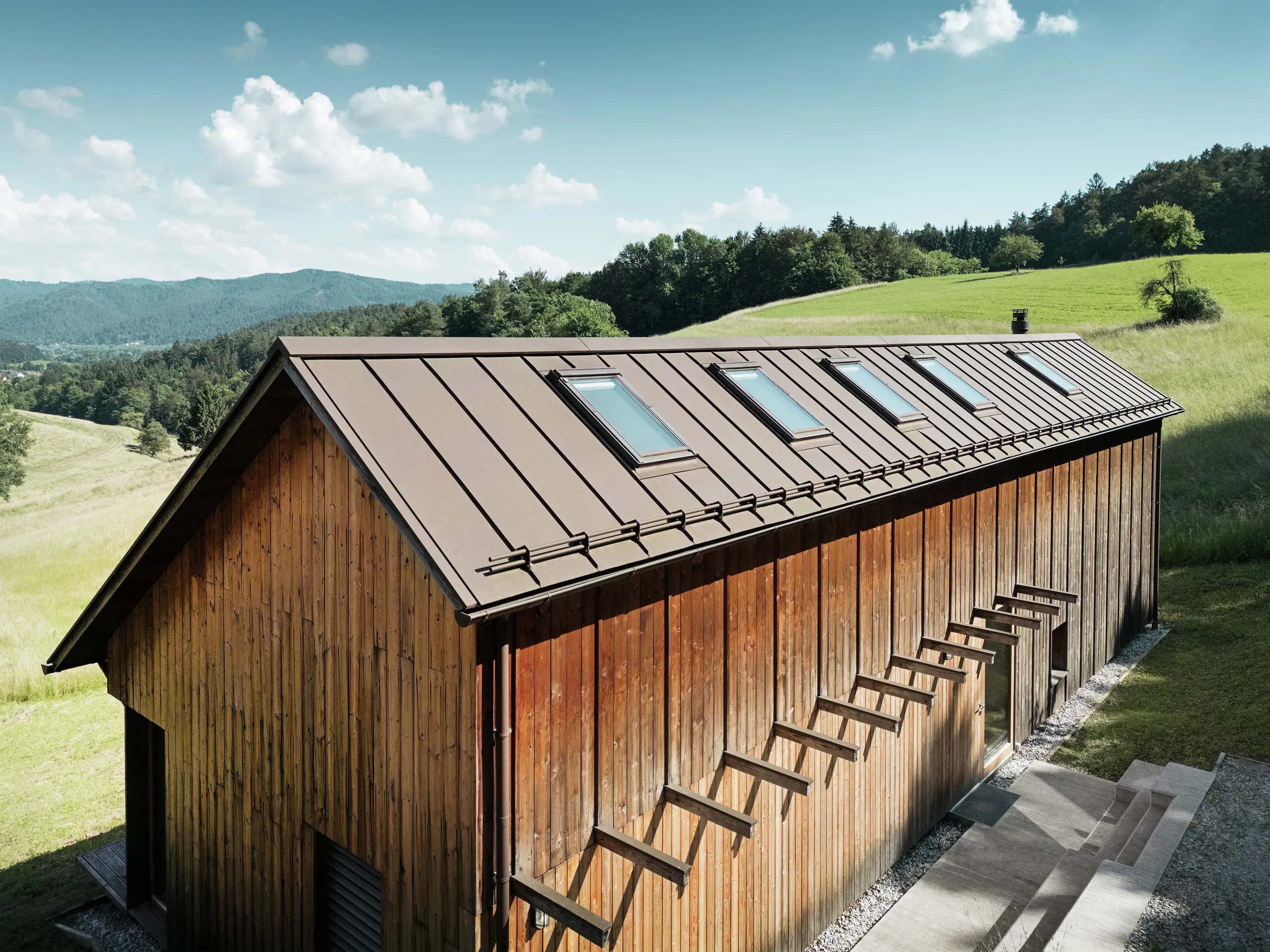 Architektonicky čistý dřevěný dům s vysoce kvalitní hliníkovou střechou PREFALZ v tmavě hnědé barvě P.10. Nenáročná střecha PREFA zaujme svými přesnými liniemi, vysokou odolností proti UV záření a korozi a perfektně integrovanými střešními okny. Kombinace udržitelné dřevěné fasády a odolné falcované střechy harmonicky zapadá do idylické kopcovité krajiny. Asymetrická sedlová střecha má pět přesně umístěných střešních oken, která zajišťují světlo do interiéru.