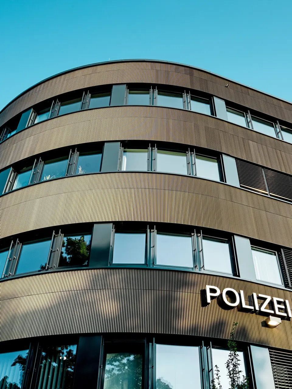 Dramatický pohled zdola na zakřivenou novostavbu policejní stanice 43 v Hamburku: Výrazná hliníková fasáda PREFA z eloxovaných extrudovaných profilů „Zacke“ ve střední bronzové barvě je architektonickým poutačem. Jemná, rytmická struktura vytváří dynamické světelné a stínové vzory a zdůrazňuje plynulé křivky budovy. Hliník PREFA nabízí vysokou odolnost proti povětrnostním vlivům, ochranu proti korozi a dlouhou životnost – udržitelné a nenáročné řešení pro precizní a moderní fasádní architekturu.