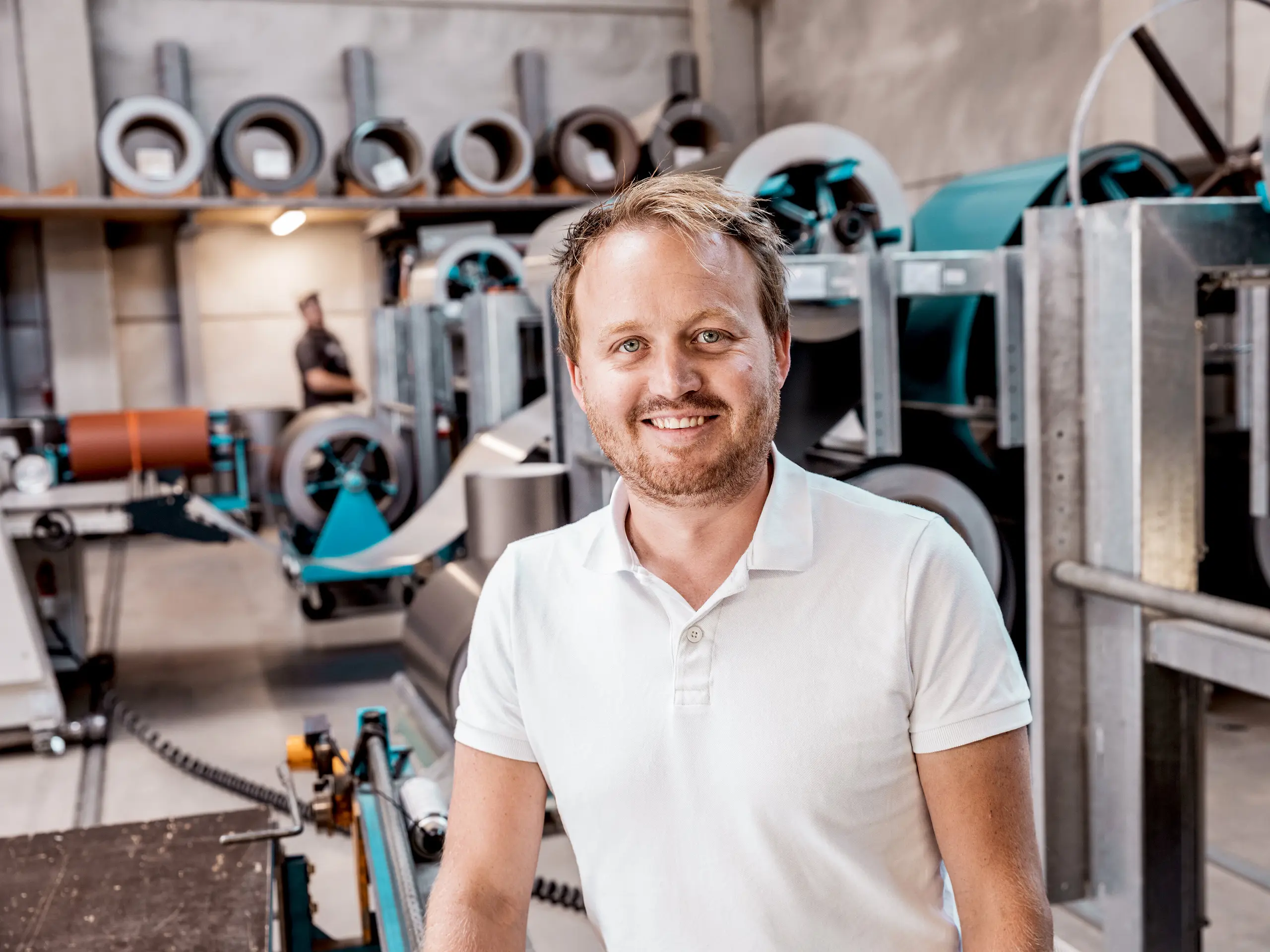 Na snímku je David Thumfart, plechář ve firmě Kapl Bau GmbH, která se zabývá pokrývačskými pracemi a plechářstvím, ve své dílně před stroji na zpracování kovových svitků. S úsměvem a profesionálním vystupováním hrdě stojí uprostřed nejmodernějšího výrobního zařízení používaného k výrobě hliníkových fasádních a střešních systémů. Stroje v pozadí ilustrují přesnost potřebnou k výrobě vysoce kvalitních produktů, jako je fasáda PREFALZ, která byla použita na rodinném domě ve Freistadtu. Thumfart tak ztělesňuje odhodlání a odborné znalosti, které se vkládají do výroby a montáže odolných a esteticky příjemných stavebních prvků.