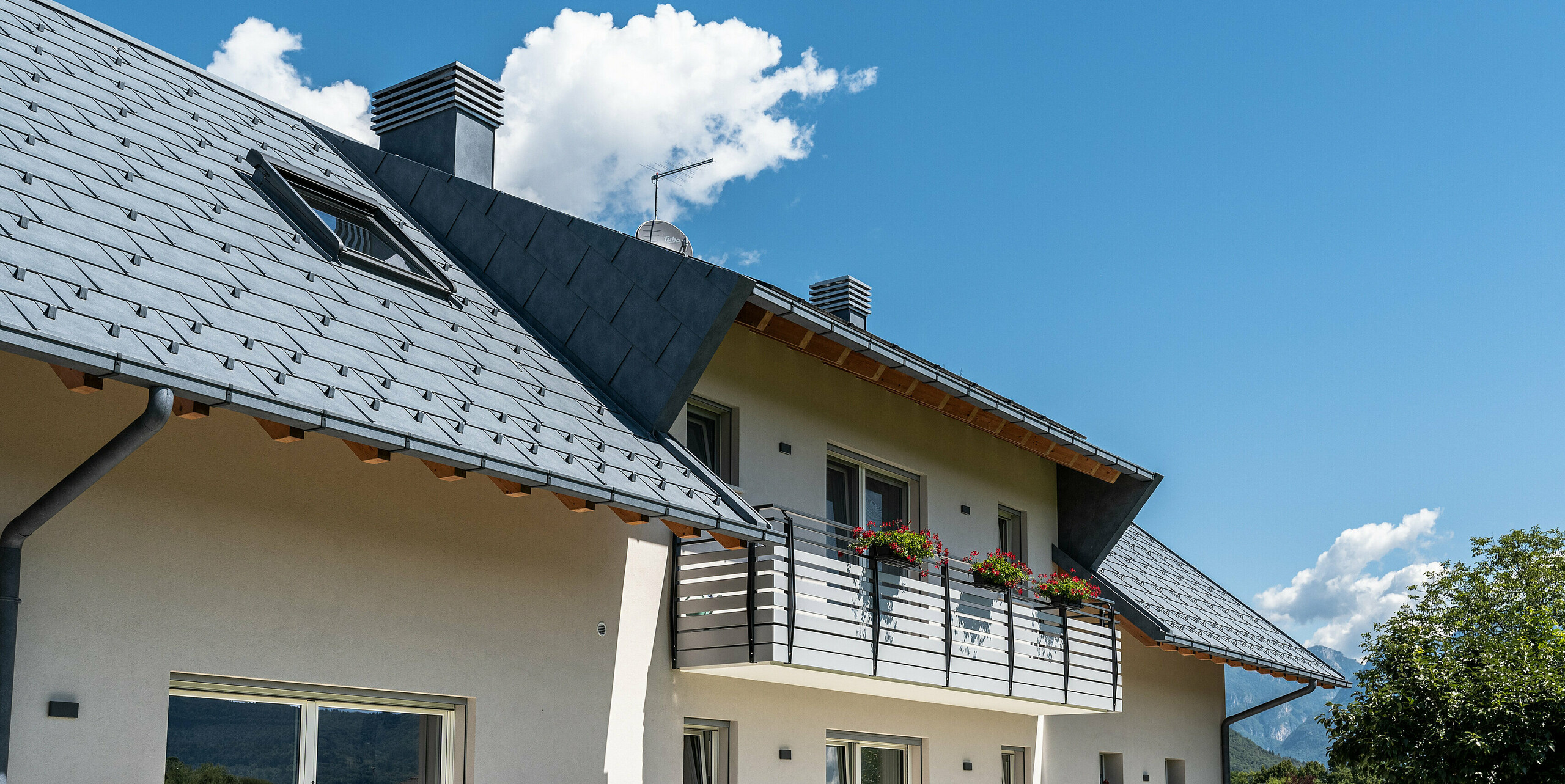 Pohled zdola na elegantní střechu rodinného domu v Belluno, Itálie. Střešní plocha má netopýří vikýř a byla pokryta střešními panely PREFA R.16 v barvě P.10 antracit. Střešní odvodnění od PREFA sestávající z krabicového žlabu a odtokové trubky je barevně sladěno s robustní hliníkovou střechou, což vytváří jednotnou a moderní estetiku a zároveň nabízí optimální ochranu.