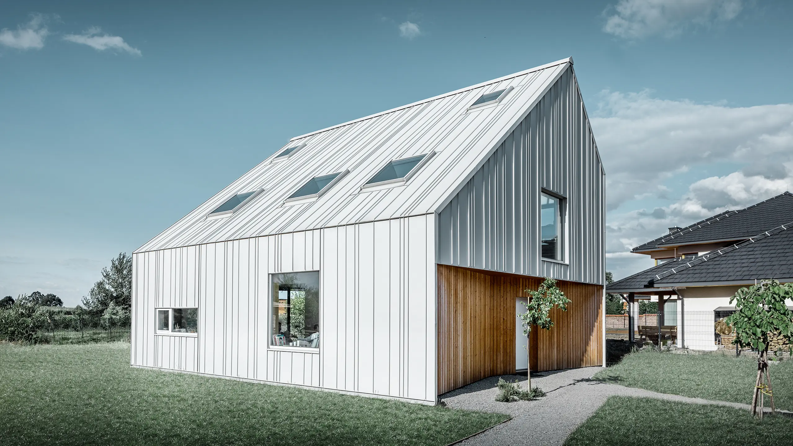 Moderně pojatý rodinný dům se sedlovou střechou, celý vnější plášť je obložen PREFALZem v barvě prefa bílá. Různě široké šáry se od střechy táhnou dále přes fasádu. Do střechy je zabudováno několik střešních oken.