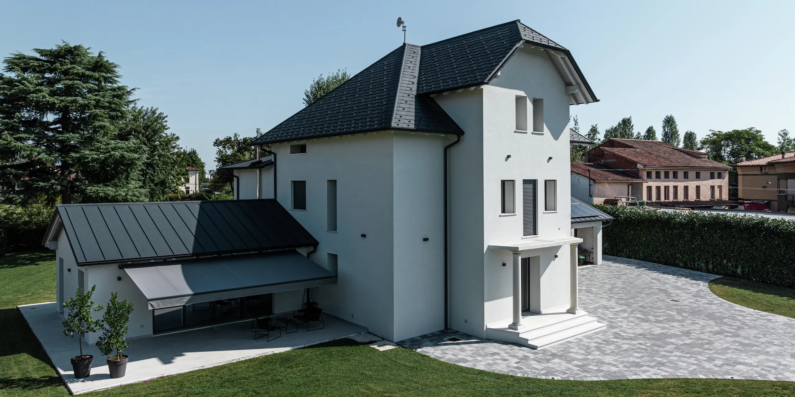 Seitenansicht eines modernen Einfamilienhauses in Marano Vicentino, Italien, mit robusten PREFA Dachschindeln in P.10 Anthrazit. Das angrenzende Nebengebäude wurde mit einer langlebigen Doppelstehfalzdeckung ausgestattet. Die hellgraue Markise spendet Schatten bei starker Mittagssonne. Die klare Architektur und die hochwertigen Aluminiumprodukte sorgen für eine moderne und witterungsbeständige Dachlandschaft.