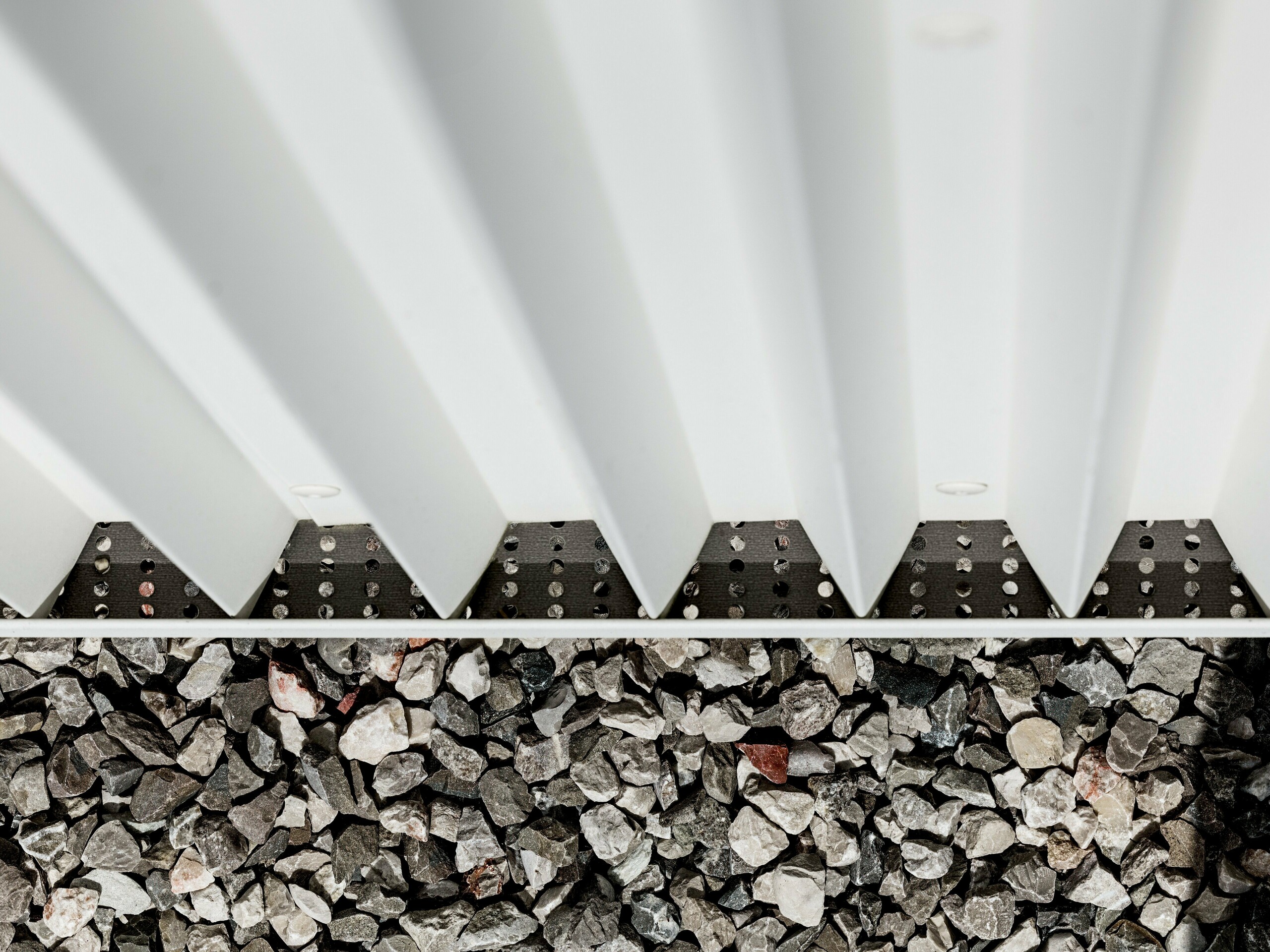 Obrázek ukazuje detail odvětrávané fasády PREFALZ z čistě bílého hliníku s přesně skládanými klikatými tvary. Tato inovativní technika skládání zajišťuje nejen vizuálně atraktivní konstrukci, ale také vynikající větrání a odolnost vůči povětrnostním vlivům. Fasádní prvky jsou spojeny pomocí specializovaných, skrytých spojovacích prvků, což vede k obzvláště čistému a odolnému povrchu. Spodní část obrázku ukazuje štěrkovou zásypku obklopující budovu, která slouží k odvodnění a stabilizaci a dále zdůrazňuje funkčnost designu fasády. Použití PREFALZ splňuje estetické i technické požadavky, což z něj činí ideální volbu pro moderní a udržitelnou architekturu.