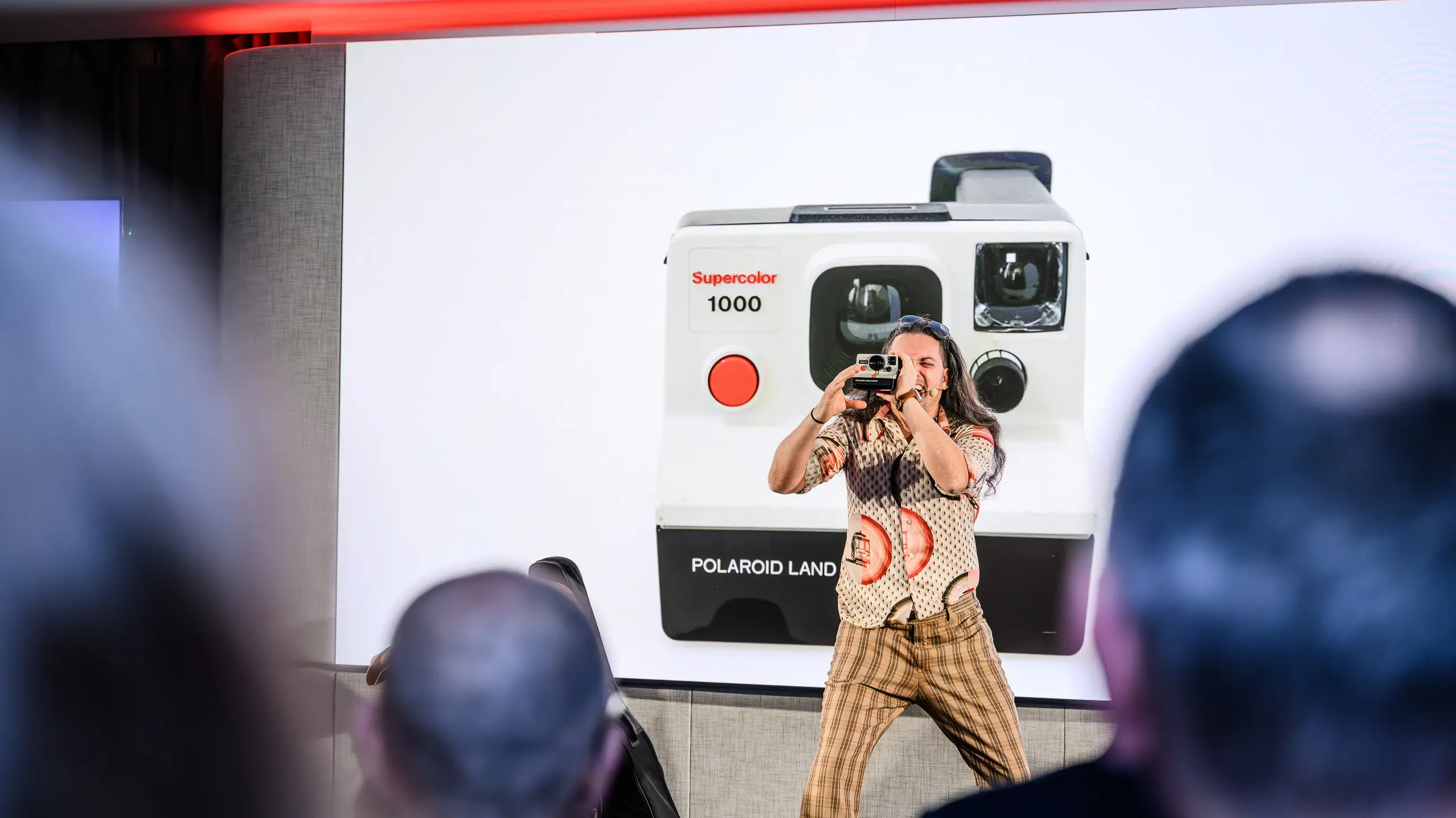 Kreativní moment na prezentaci PREFARENZEN 2026: Zábavný moderátor zachycuje publikum retro fotoaparátem Polaroid – před nadrozměrnou projekcí ikonického Supercolor 1000. Tento umělecký přínos překlenuje propast mezi minulostí a budoucností, stejně jako PREFA se svými odolnými hliníkovými výrobky. Večer plný emocí, inovací a estetické inspirace.