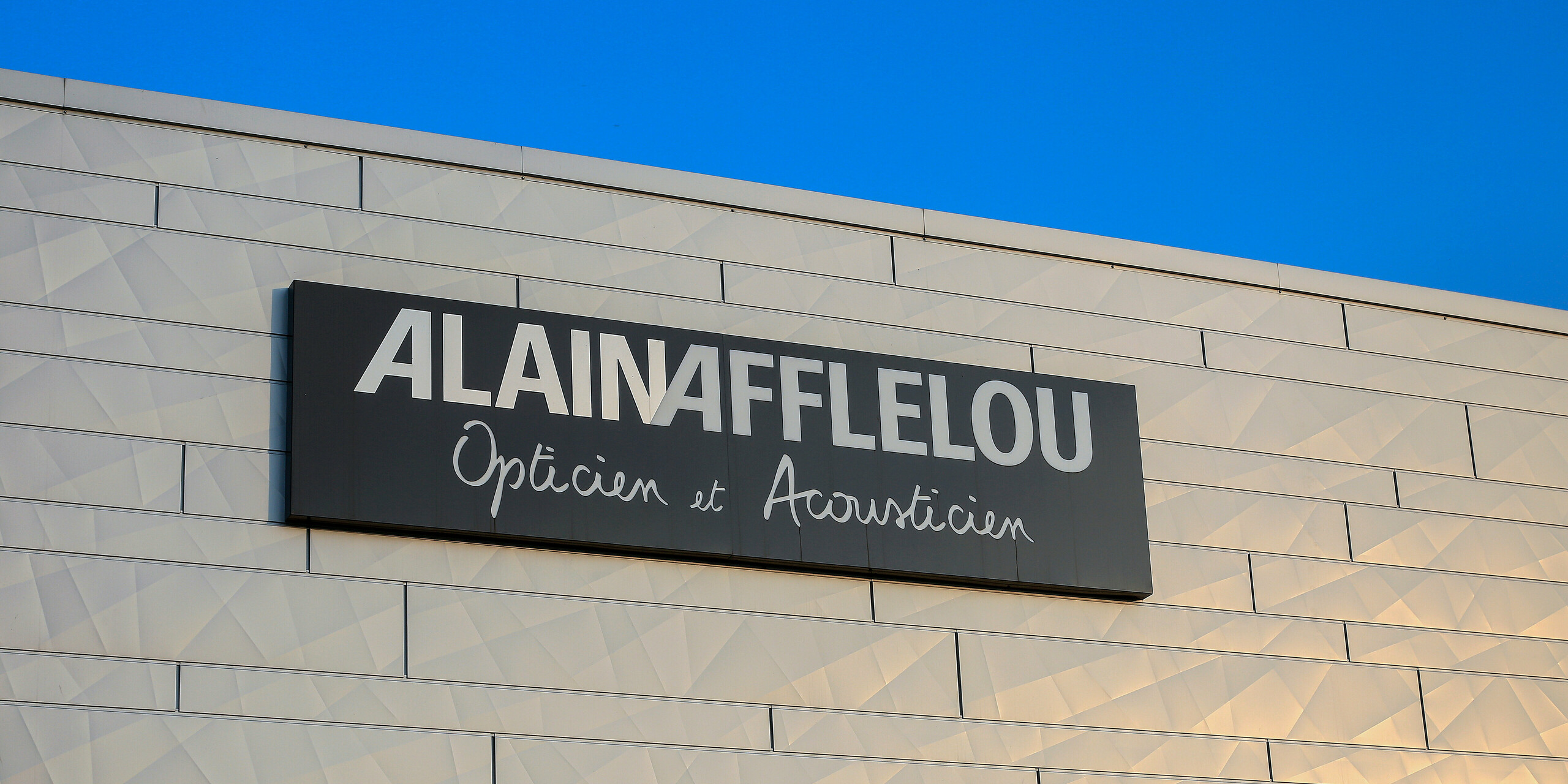 Detailní záběr na nápis společnosti AFFLELOU na budově obchodu s optikou a akustikou v Auray - kolem loga jsou vidět třpytivé stříbrné fasádní lamely PREFA Siding.X.