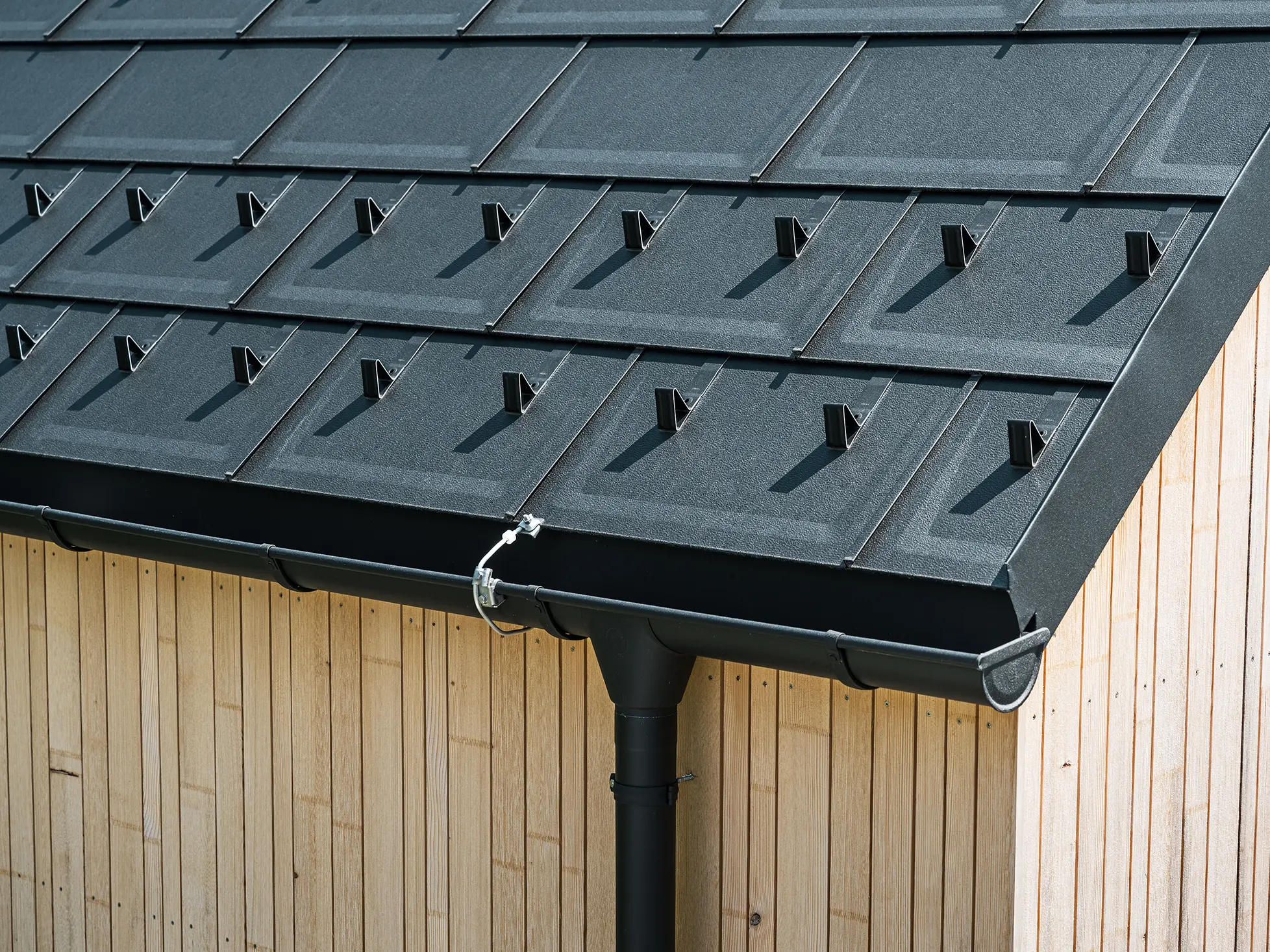 Detailní záběr PREFA střešních panelů R.16 se sněhovými háky, střešním žlabem a kruhovým svodem v provedení P.10 černá, včetně hromosvodu a svislé dřevěné fasády