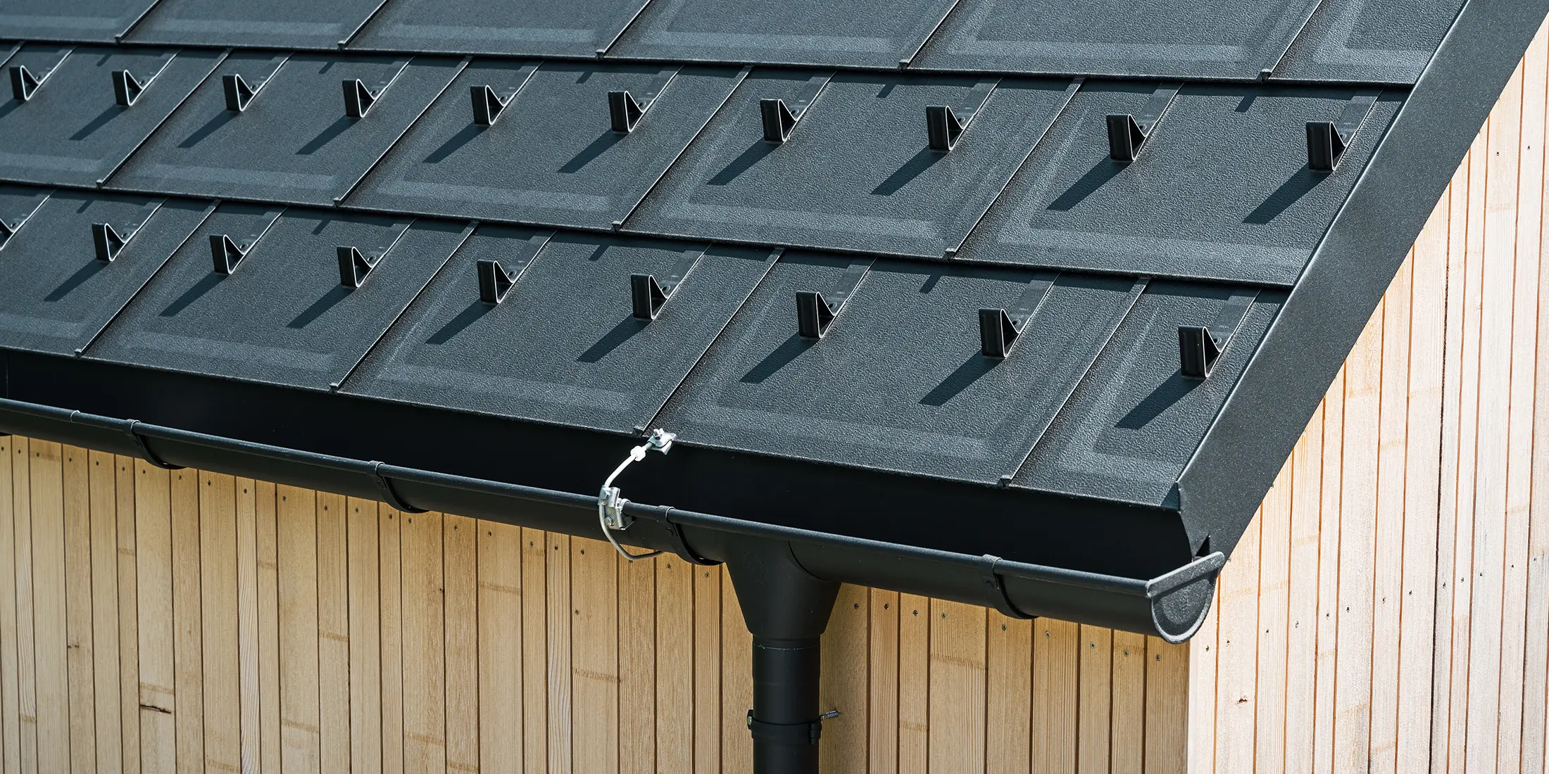 Detailní záběr PREFA střešních panelů R.16 se sněhovými háky, střešním žlabem a kruhovým svodem v provedení P.10 černá, včetně hromosvodu a svislé dřevěné fasády