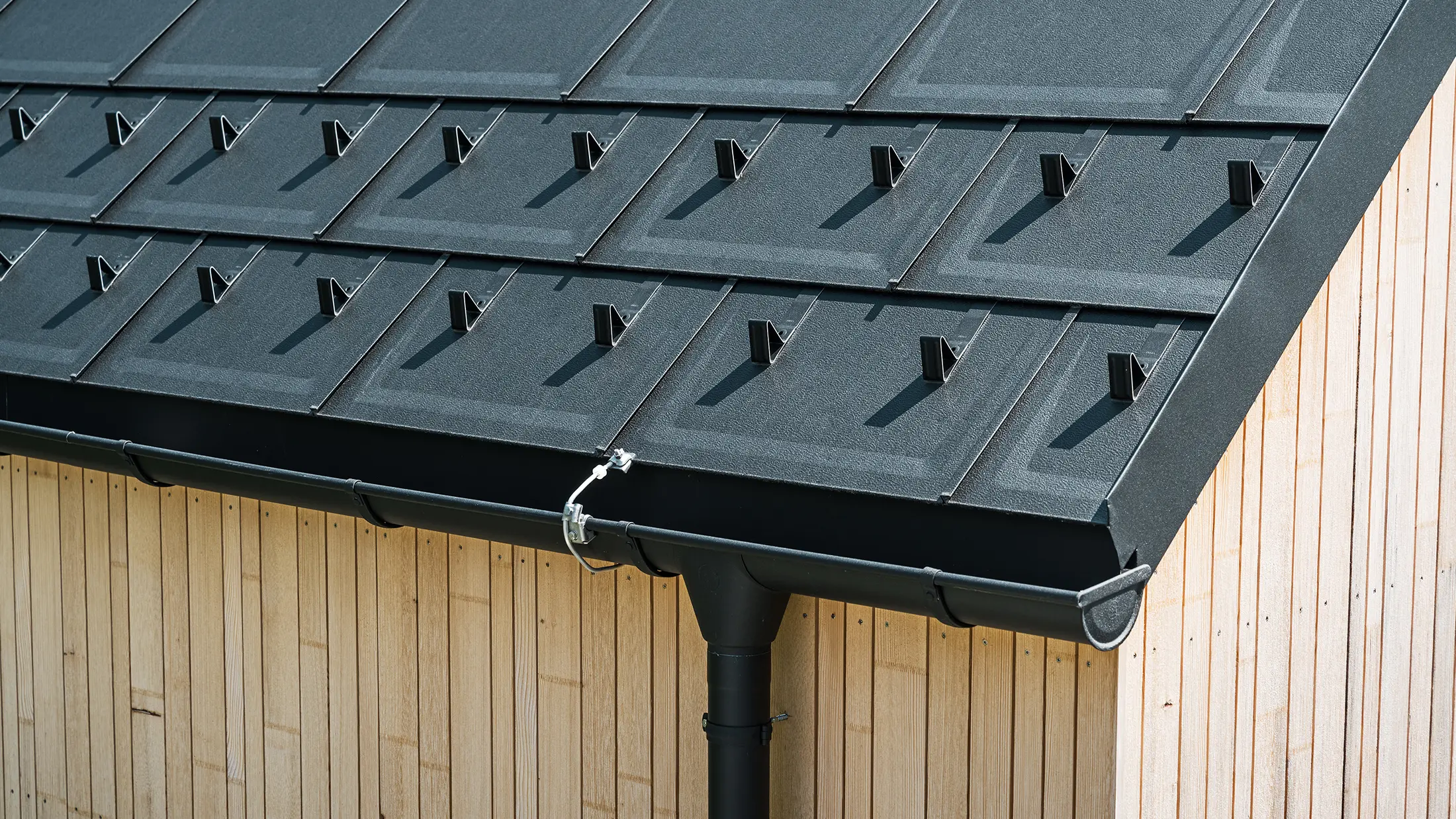 Detailní záběr PREFA střešních panelů R.16 se sněhovými háky, střešním žlabem a kruhovým svodem v provedení P.10 černá, včetně hromosvodu a svislé dřevěné fasády