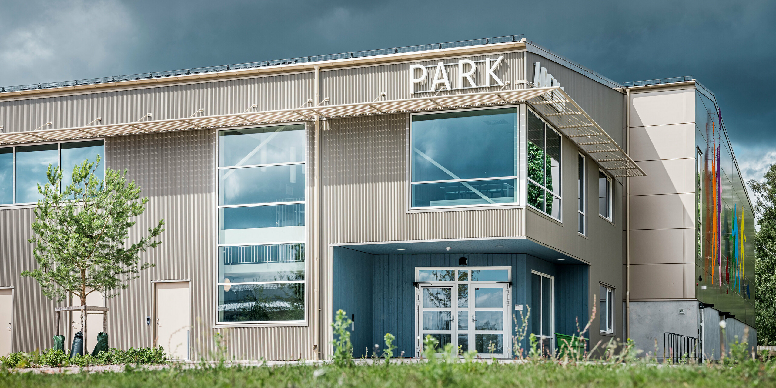 Vstupní prostor Park Areny ve Värnamu ve Švédsku. Fasáda je obložena výrazným pilovým Zackenprofilem PREFA v bronzové barvě, který vytváří hru světel a stínů. Moderní prosklená čela a umělecky řešené vyobrazení sportovců na plášti budovy zdůrazňují inovativní design sportovní haly.