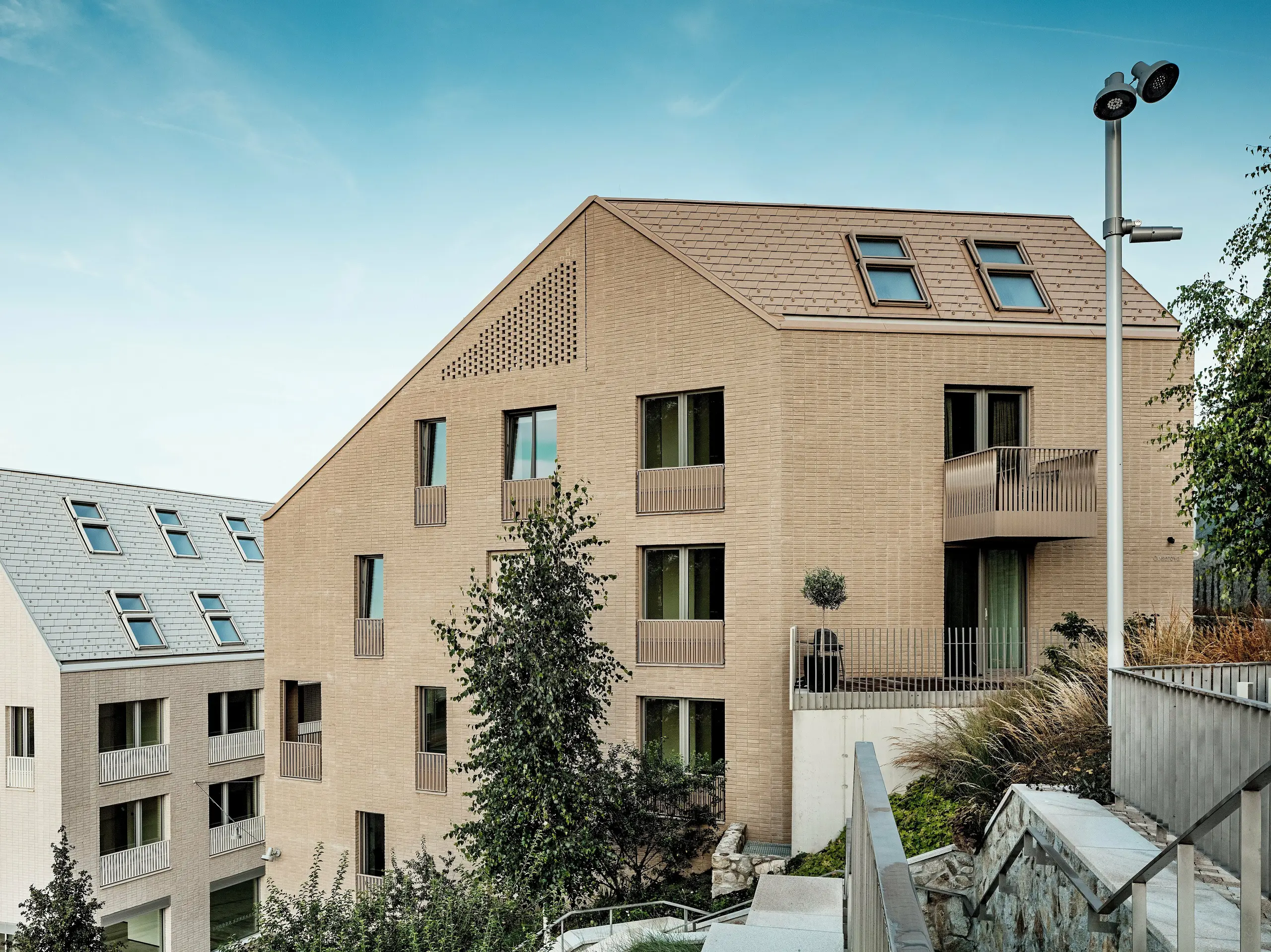 Moderní obytné budovy v bratislavské čtvrti Vydrica se vyznačují jasným, minimalistickým designovým jazykem a harmonickým začleněním do terénu: Strmé šikmé střechy s přesně položenými hliníkovými šindeli PREFA v barvách P.10 pískově hnědá a prefa bílá vytvářejí svými texturovanými povrchy a integrovanými střešními okny výrazné vizuální akcenty. Jednotný design střechy a fasády vytváří klidné, vysoce kvalitní povrchy. Hliník PREFA přesvědčuje svou trvanlivostí, snadnou montáží, vysokou odolností vůči povětrnostním vlivům a umožňuje sofistikovaná a udržitelná střešní řešení.