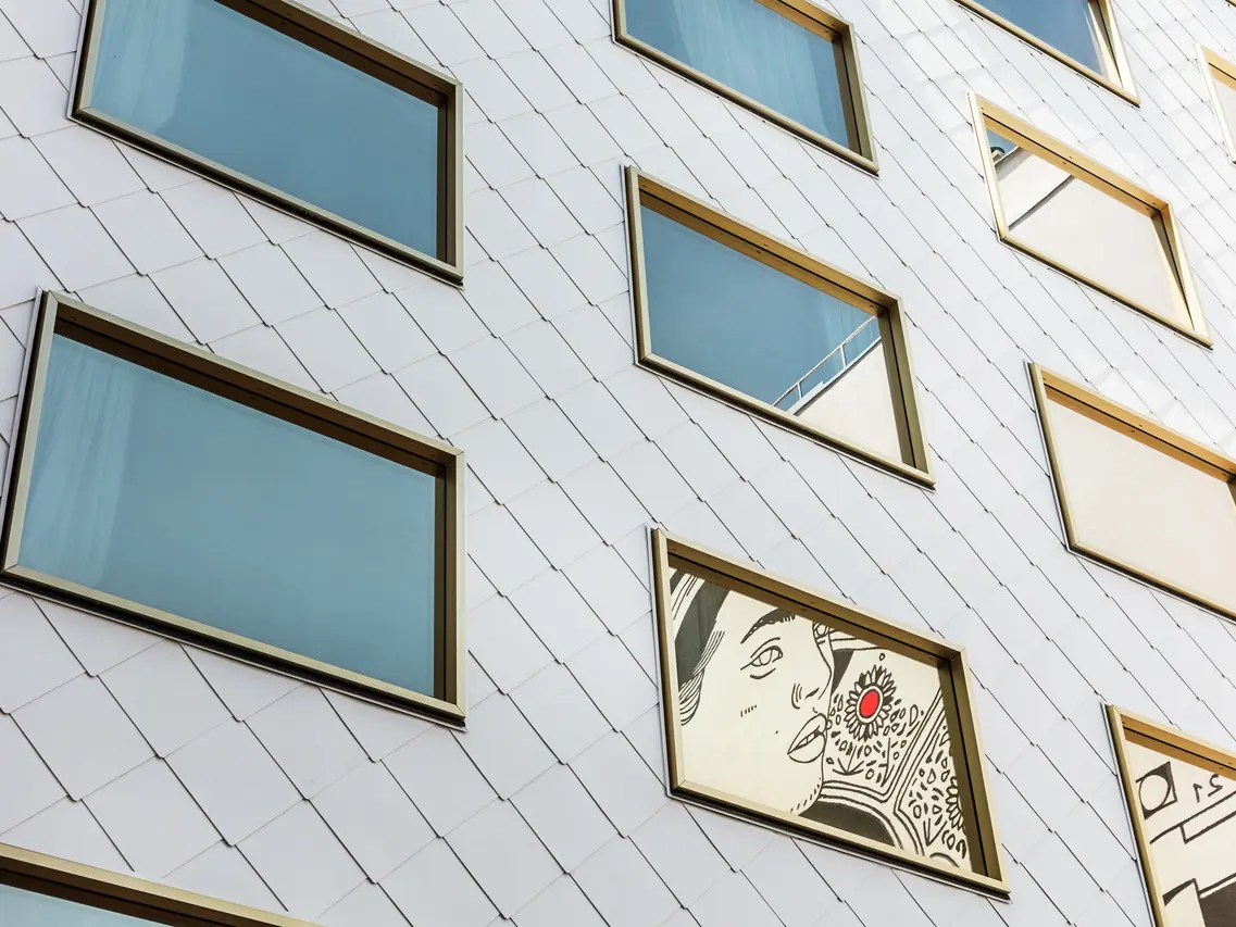 Detailní pohled na fasádu hotelu „THE ROCK Radisson RED Vienna“, která je obložena PREFA hliníkovými šablonami PREFA 44 × 44 v barvě P.10 čistě bílé. Mezi několika zrcadlově zasklenými okny se zlatými rámy vyniká okno, ve kterém je zasazeno umělecké dílo zobrazující portrét osoby s výrazným červeným akcentem.