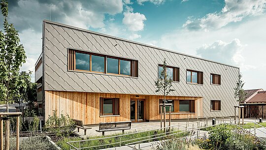 Čelní pohled na školku v Alzenau ukazuje lákavý hlavní vchod s harmonickou kombinací bronzově zbarvených stěnových panelů PREFA (44 x 44 cm) a vertikální dřevěné fasády v přízemí. Jasně strukturovaný hliníkový obklad vytváří elegantní šupinovitý vzor, ​​rytmicky přerušovaný velkorysými okny s dřevěnými rámy a vertikálními žaluziemi. V popředí zdůrazňují bezbariérové ​​chodníky, květinové záhony a lavičky vysokou kvalitu prostoru. Souhra odolného hliníku, přírodního dřeva a promyšlené úpravy terénu činí z budovy architektonickou přehlídku udržitelných, funkčních a esteticky příjemných vzdělávacích budov s fasádními systémy PREFA.