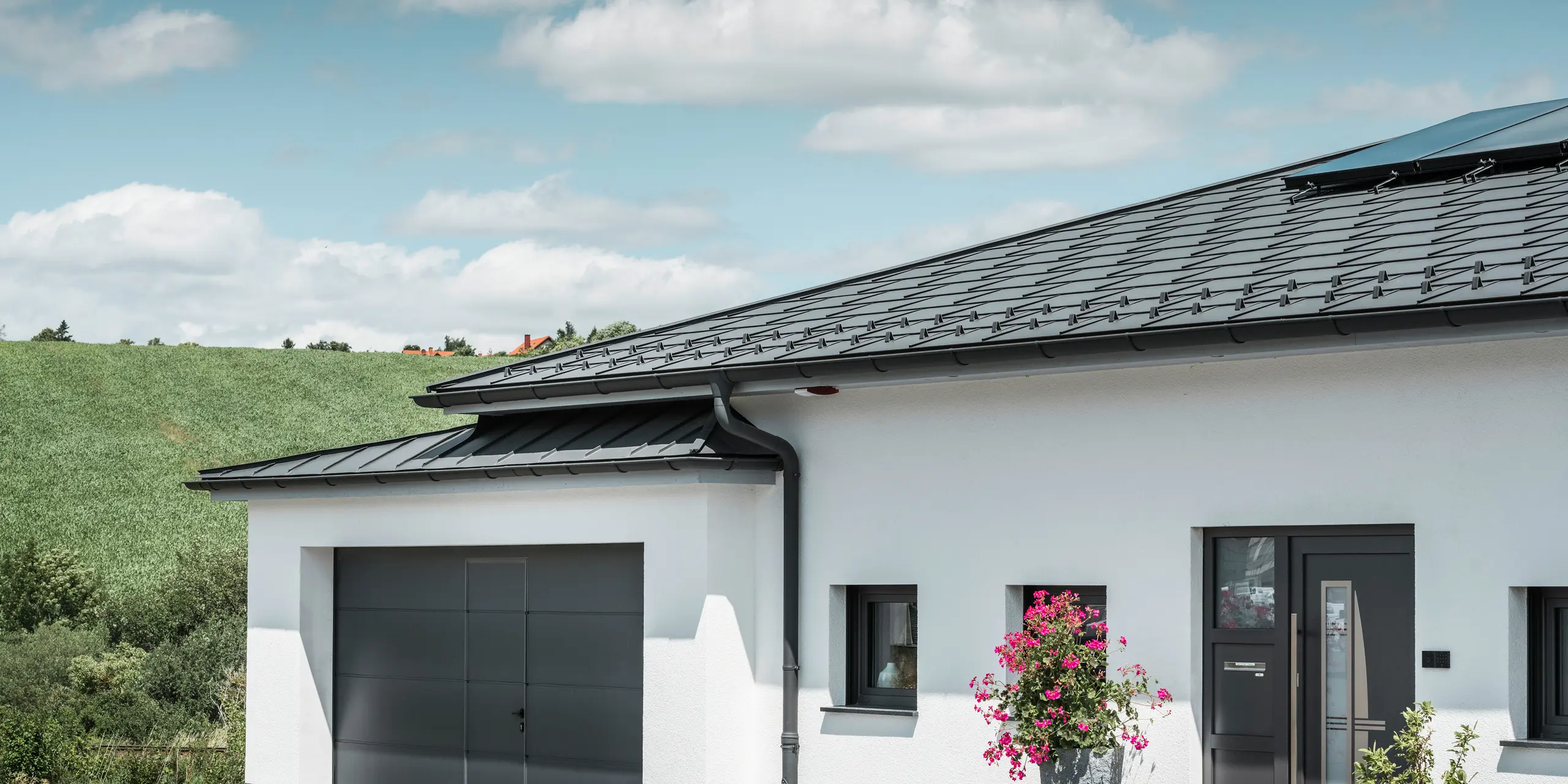 Obrázek představuje přední stranu rodinného domu v Ilmenau v Durynsku s elegantní střechou PREFA v provedení P.10 tmavě šedá. Střešní panel R.16 dodává domu moderní vzhled, zatímco integrovaný okapový systém PREFA s tmavě šedým půlkulatým žlabem a kruhovým svodem zajišťuje účinný odvod dešťové vody. Garáž, vybavená tmavě šedým PREFALZem, harmonicky zapadá do celkové estetiky a podtrhuje kvalitu a styl hliníkových produktů PREFA.