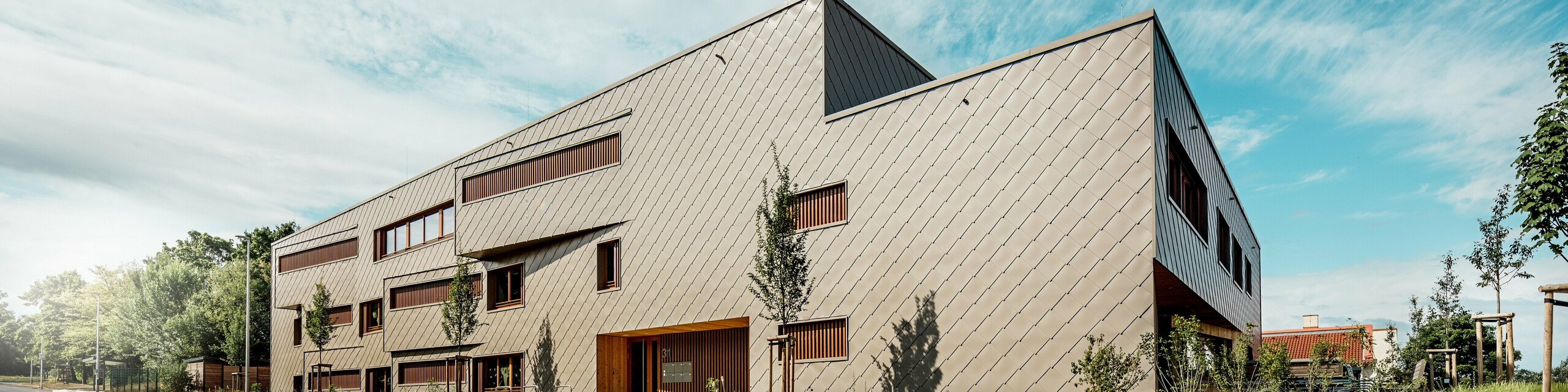 Při pohledu z úhlu pohledu se školka Alzenau prezentuje svými výraznými, dynamickými liniemi a vysoce kvalitní hliníkovou fasádou PREFA v bronzové barvě. Diagonální uspořádání fasádních šablon o rozměrech 44 x 44 cm dodává budově jasnou strukturu a elegantní, homogenní povrch. Schůdkové okenní otvory a vertikální dřevěné žaluzie vytvářejí rytmické akcenty, zatímco minimalistický designový jazyk vyjadřuje moderní zdrženlivost. Kombinace odolného hliníku, přírodního dřeva a promyšleného konceptu zeleně podél ulice podtrhuje vysoké standardy udržitelnosti a kvality výstavby. Tento snímek působivě ukazuje, jak může být funkční architektura s produkty PREFA esteticky působivá a stanovovat standardy urbanistického plánování.