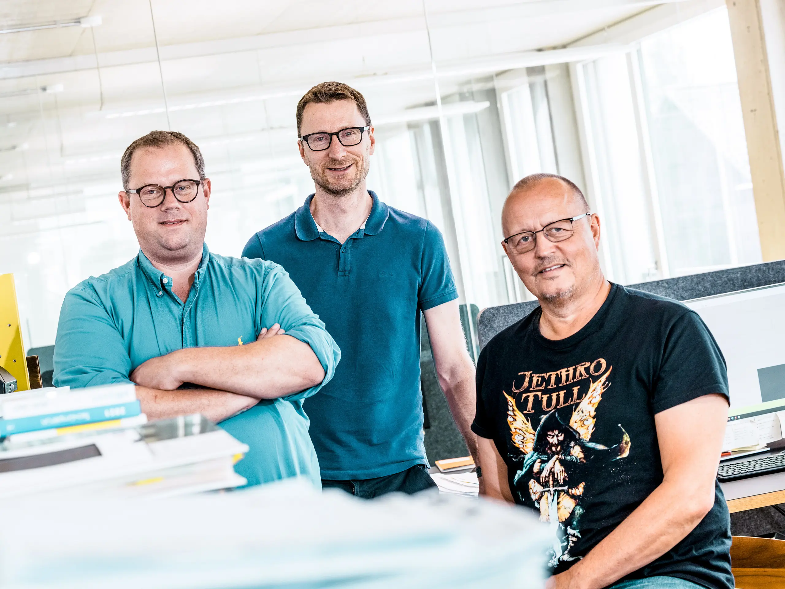 Týmová fotografie architektů z Kaundbe v jejich moderní kanceláři: Tři architekti stojí sebevědomě před svými pracovišti a technickou dokumentací. V popředí jsou hromady plánů a složek s projekty, zatímco v pozadí jsou vidět počítačové obrazovky zobrazující stavební výkresy. Scéna vyjadřuje kompetenci, spolupráci a technickou přesnost – klíčové hodnoty při plánování a realizaci fasádních řešení s hliníkovými výrobky PREFA.