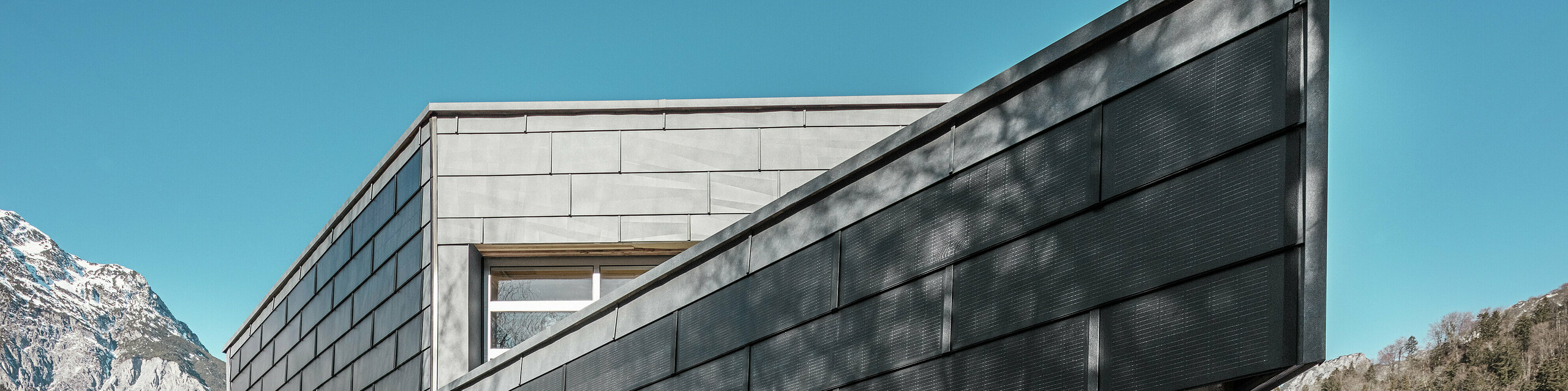 Budova kovovýrobní společnosti RÜMA ve Vorarlbersku zaujme svou moderní fasádou ze solárních střešních panelů PREFA a pohledového betonu. Obklopena horami a stromy, jejichž stíny vrhají na zeď, stojí pod jasnou modrou oblohou. Špičatý roh budovy a fasádní panely PREFA FX.12 dále zdůrazňují její výrazný vzhled.