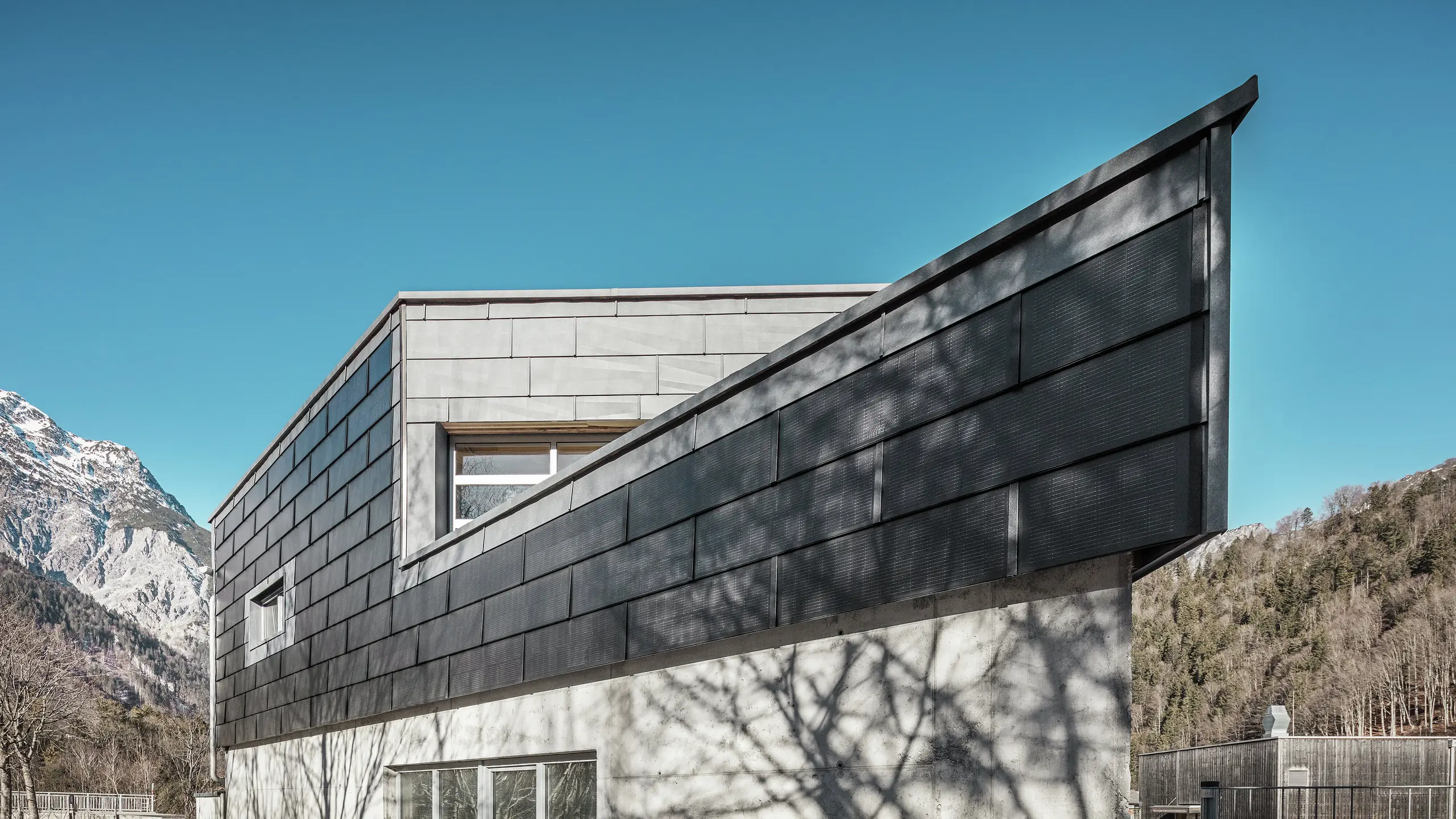 Budova kovovýrobní společnosti RÜMA ve Vorarlbersku zaujme svou moderní fasádou ze solárních střešních panelů PREFA a pohledového betonu. Obklopena horami a stromy, jejichž stíny vrhají na zeď, stojí pod jasnou modrou oblohou. Špičatý roh budovy a fasádní panely PREFA FX.12 dále zdůrazňují její výrazný vzhled.