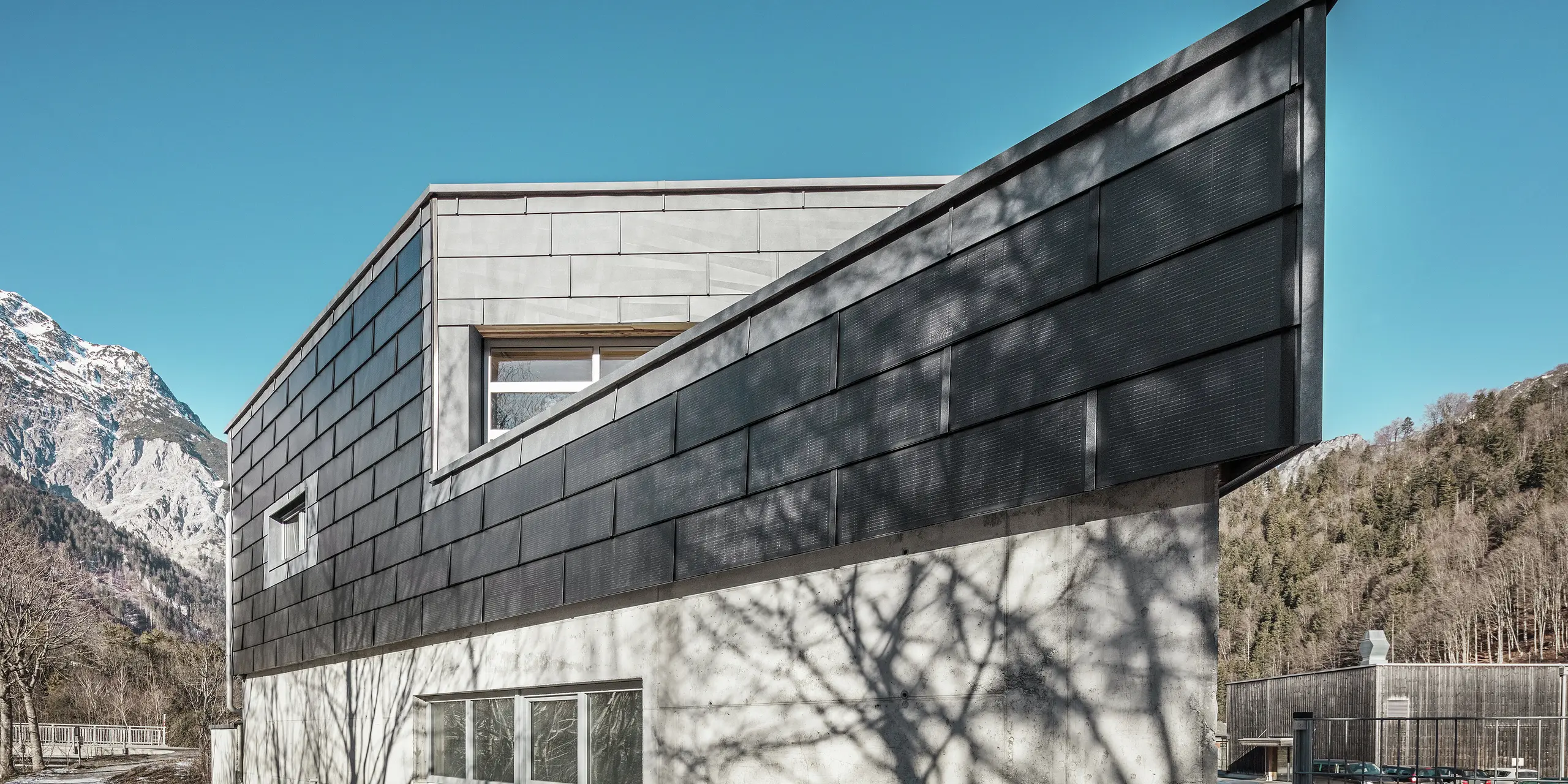 Budova kovovýrobní společnosti RÜMA ve Vorarlbersku zaujme svou moderní fasádou ze solárních střešních panelů PREFA a pohledového betonu. Obklopena horami a stromy, jejichž stíny vrhají na zeď, stojí pod jasnou modrou oblohou. Špičatý roh budovy a fasádní panely PREFA FX.12 dále zdůrazňují její výrazný vzhled.
