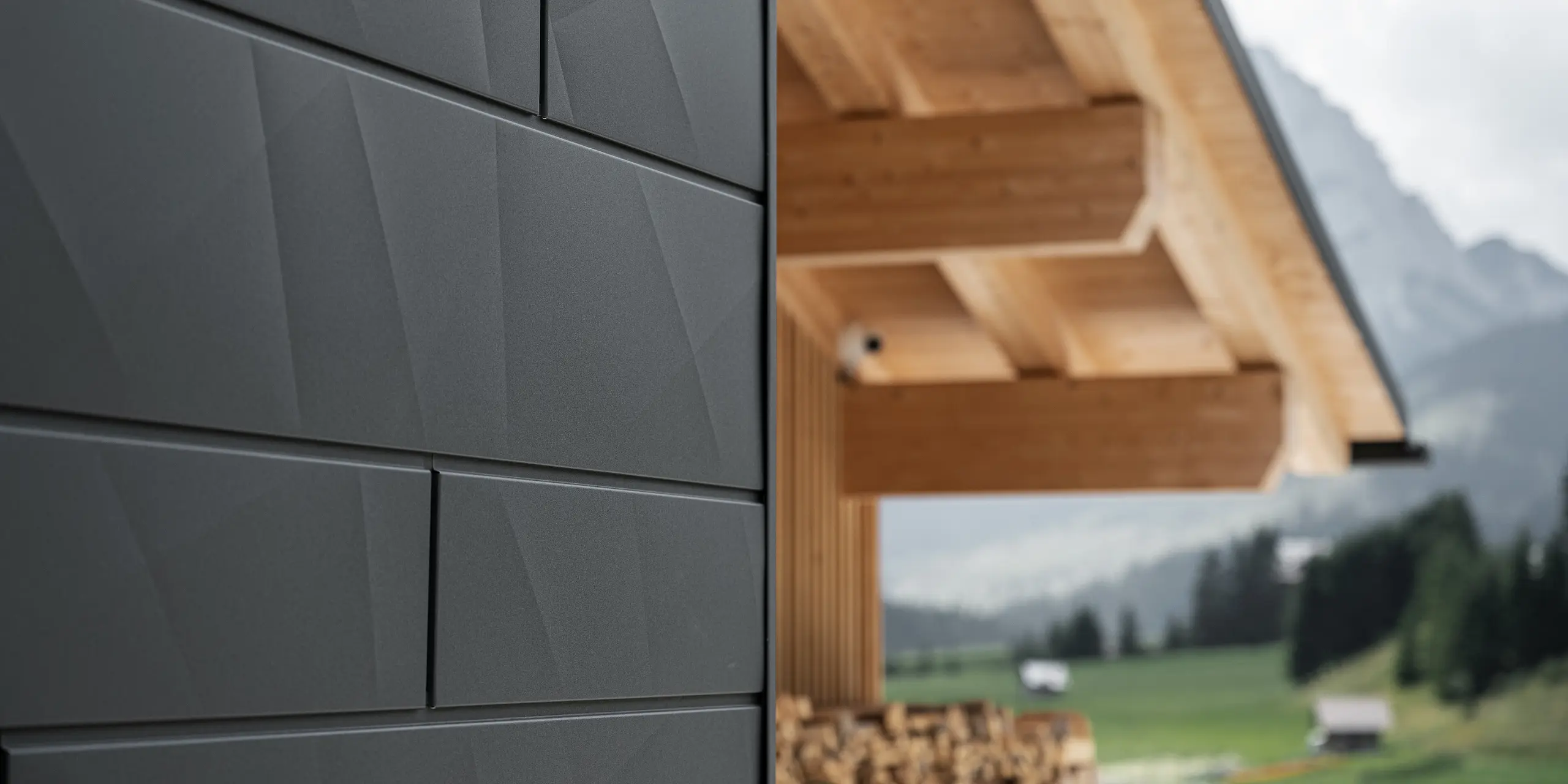 Detailní záběr fasádních lamel Siding.X v provedení P.10 černá od společnosti PREFA na budově Casa Hoffe v italské obci Sappada. Hliníkový fasádní systém je mistrně proveden v moderním abstraktním designu, který vytváří působivý kontrast s viditelnými dřevěnými prvky krovu. Toto inovativní řešení fasády se dokonale začleňuje do alpského prostředí a představuje spojení funkčnosti, trvanlivosti a estetické architektury.