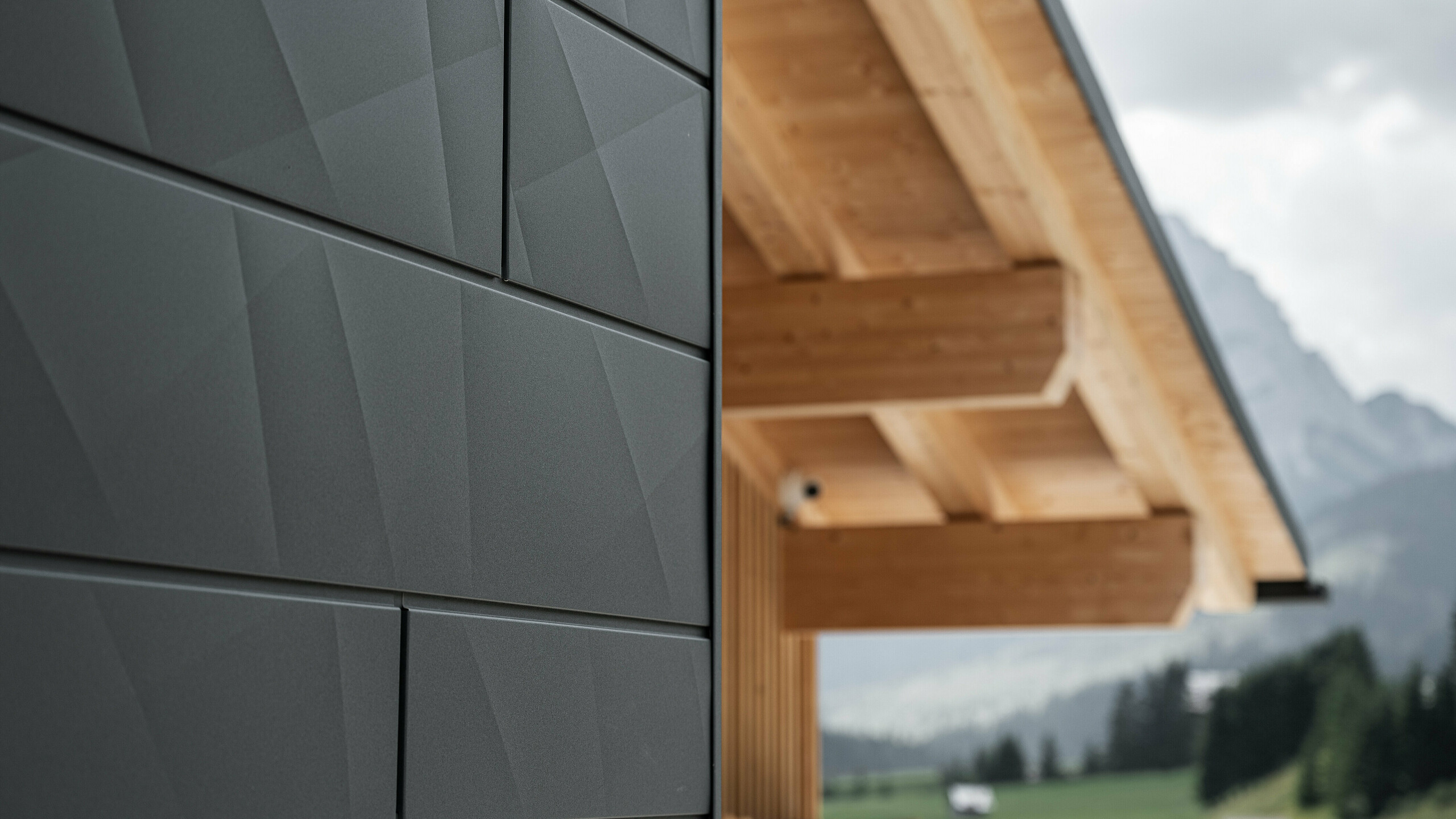 Detailní záběr fasádních panelů Siding.X v černé barvě P.10 od PREFA na Casa Hoffe v italském Sappadě. Hliníkový fasádní systém je mistrně proveden v moderním abstraktním designu, který vytváří atraktivní kontrast k viditelným dřevěným prvkům krovu. Inovativní fasádní řešení se dokonale začleňuje do alpského prostředí a představuje spojení funkčnosti, odolnosti a estetické stavební kultury.