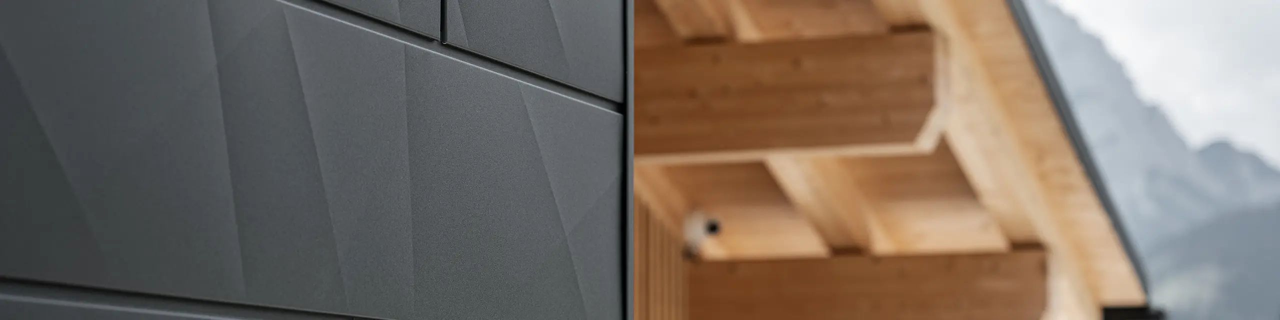Detailní záběr fasádních lamel Siding.X v provedení P.10 černá od společnosti PREFA na budově Casa Hoffe v italské obci Sappada. Hliníkový fasádní systém je mistrně proveden v moderním abstraktním designu, který vytváří působivý kontrast s viditelnými dřevěnými prvky krovu. Toto inovativní řešení fasády se dokonale začleňuje do alpského prostředí a představuje spojení funkčnosti, trvanlivosti a estetické architektury.