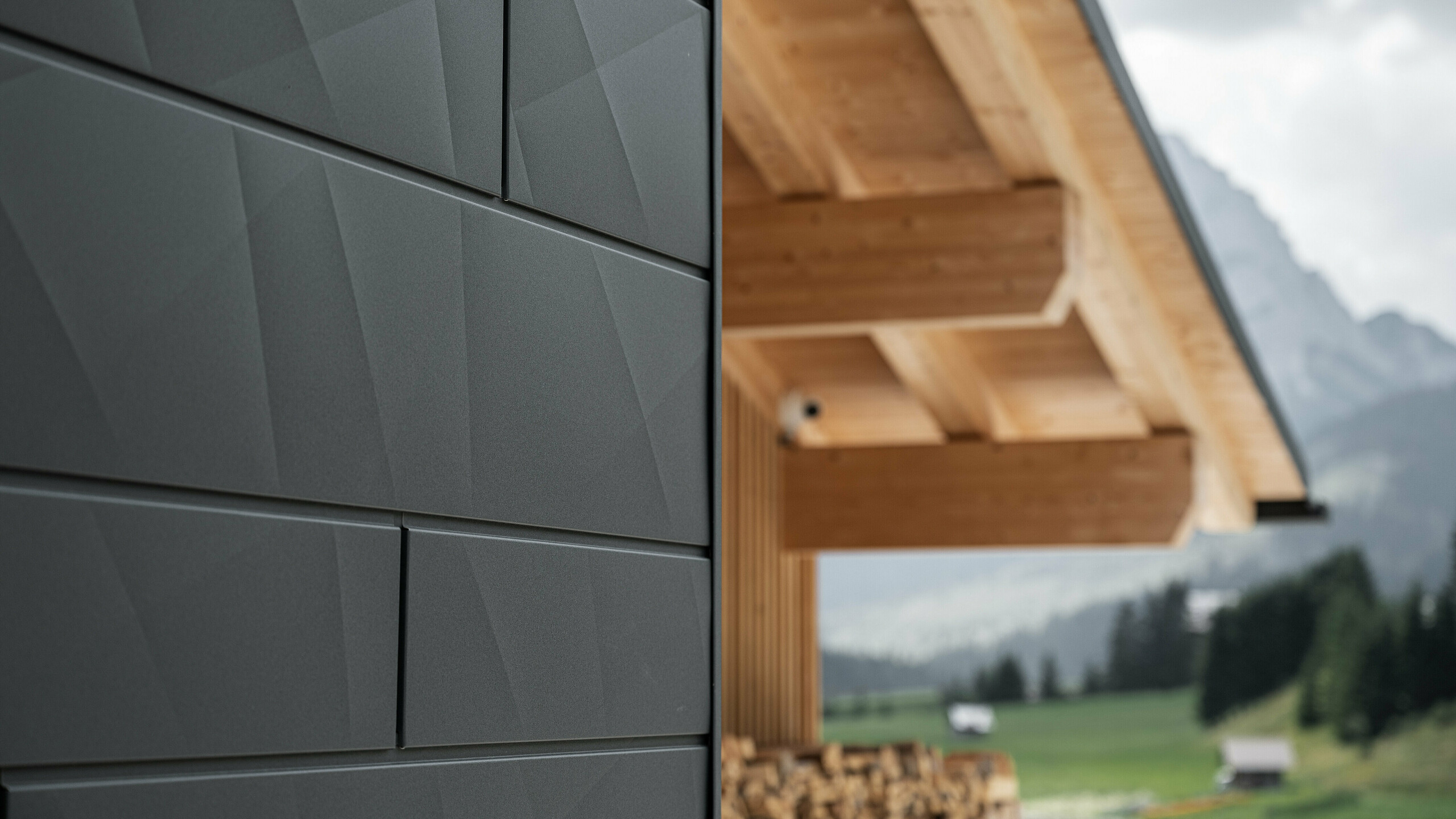 Detailní záběr fasádních panelů Siding.X v černé barvě P.10 od PREFA na Casa Hoffe v italském Sappadě. Hliníkový fasádní systém je mistrně proveden v moderním abstraktním designu, který vytváří atraktivní kontrast k viditelným dřevěným prvkům krovu. Inovativní fasádní řešení se dokonale začleňuje do alpského prostředí a představuje spojení funkčnosti, odolnosti a estetické stavební kultury.