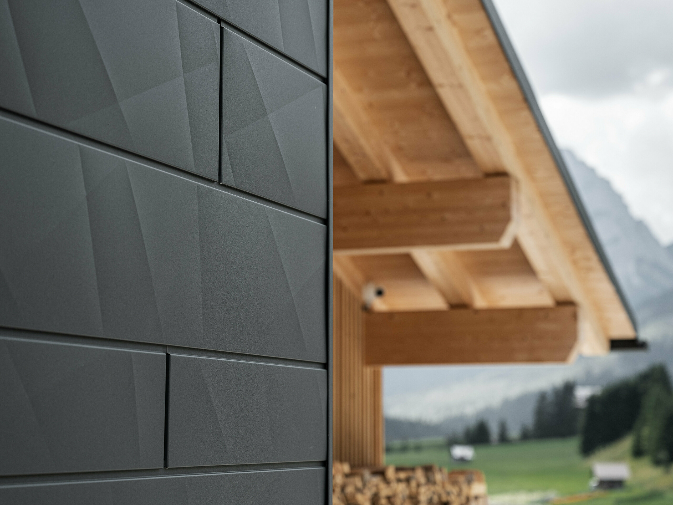Detailní záběr fasádních lamel Siding.X v provedení P.10 černá od společnosti PREFA na budově Casa Hoffe v italské obci Sappada. Hliníkový fasádní systém je mistrně proveden v moderním abstraktním designu, který vytváří působivý kontrast s viditelnými dřevěnými prvky krovu. Toto inovativní řešení fasády se dokonale začleňuje do alpského prostředí a představuje spojení funkčnosti, trvanlivosti a estetické architektury.