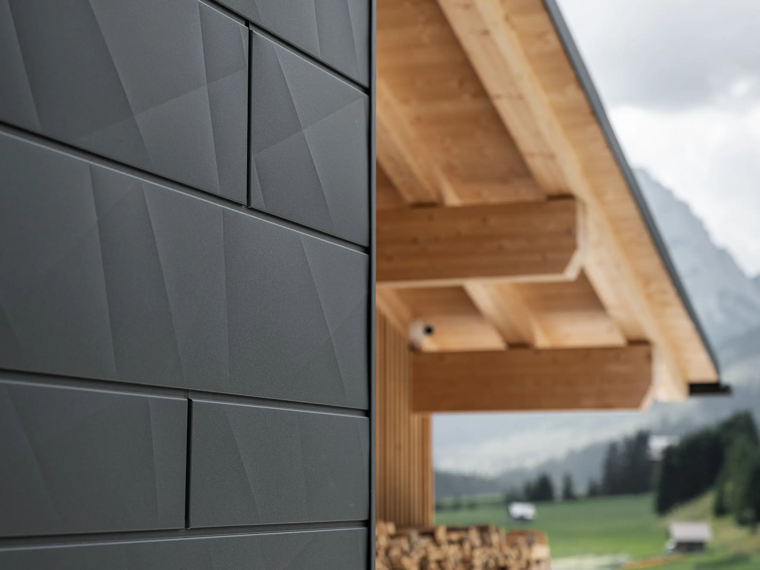 Detailní záběr fasádních lamel Siding.X v provedení P.10 černá od společnosti PREFA na budově Casa Hoffe v italské obci Sappada. Hliníkový fasádní systém je mistrně proveden v moderním abstraktním designu, který vytváří působivý kontrast s viditelnými dřevěnými prvky krovu. Toto inovativní řešení fasády se dokonale začleňuje do alpského prostředí a představuje spojení funkčnosti, trvanlivosti a estetické architektury.