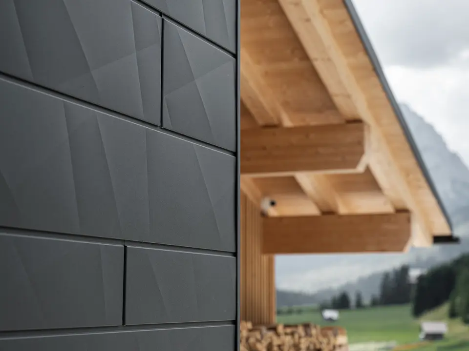 Detailní záběr fasádních lamel Siding.X v provedení P.10 černá od společnosti PREFA na budově Casa Hoffe v italské obci Sappada. Hliníkový fasádní systém je mistrně proveden v moderním abstraktním designu, který vytváří působivý kontrast s viditelnými dřevěnými prvky krovu. Toto inovativní řešení fasády se dokonale začleňuje do alpského prostředí a představuje spojení funkčnosti, trvanlivosti a estetické architektury.