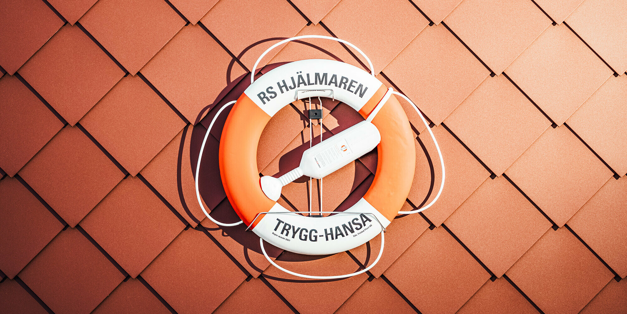Detailní záběr na fasádu domu vodní záchranné stanice na jezeře Hjälmaren ve Švédsku s jasně oranžovým záchranným kruhem na zdi. Záchranný kruh nese nápis „RS HJÄLMAREN TRYGG-HANSA“, orámovaný charakteristickými fasádními šablonami PREFA 29×29 v barvě P.10 tmavě červené, jejichž jednotná struktura vyjadřuje sílu a spolehlivost. Záchranný kruh, symbol bezpečí na vodě, nápadně kontrastuje se strukturovaným červeným hliníkovým povrchem a podtrhuje odolnost budovy vůči drsným švédským povětrnostním podmínkám.