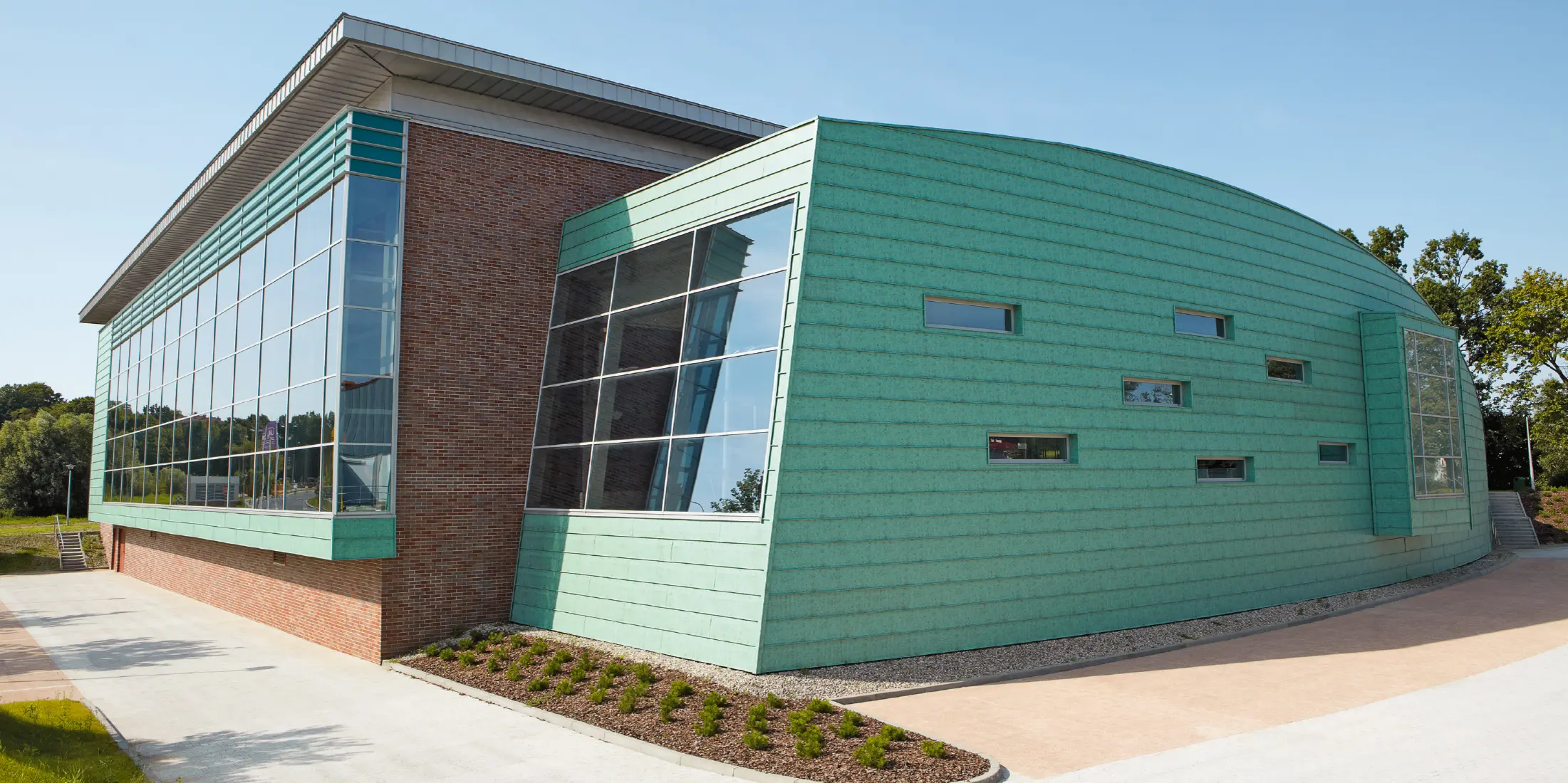 Modernes Schwimmbadgebäude mit auffälliger Fassade aus patinagrünen Aluminium-Paneelen und großen Glasflächen. Der Bau kombiniert gebogene und kantige Formen sowie unterschiedliche Materialien wie Metall und Backstein. Kleine, schmale Fenster lockern die Fassade auf. Das Gebäude steht in einer gepflegten Umgebung unter klarem, blauem Himmel.