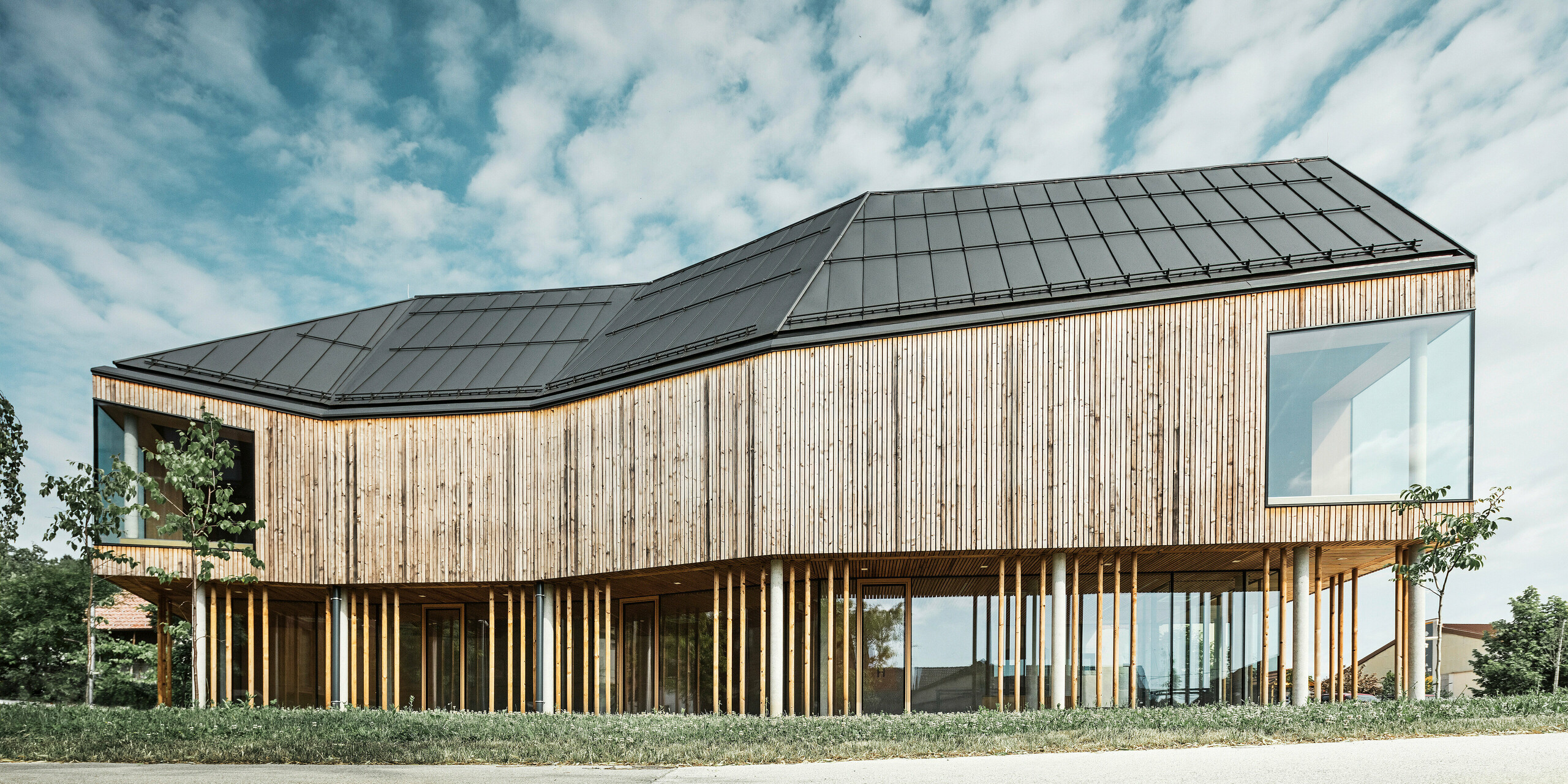 Boční pohled na ekologicky navržené návštěvnické centrum v Ig ve Slovinsku s tmavě šedou střechou PREFALZ, která zdůrazňuje čisté linie budovy a moderní architektonický jazyk. Vzájemné působení přírodního dřevěného obkladu a rozsáhlého prosklení, které odráží modrou oblohu a mraky, demonstruje spojení funkčnosti a estetiky. Design dokonale zapadá do venkovského prostředí a odráží inovativní stavební postupy.