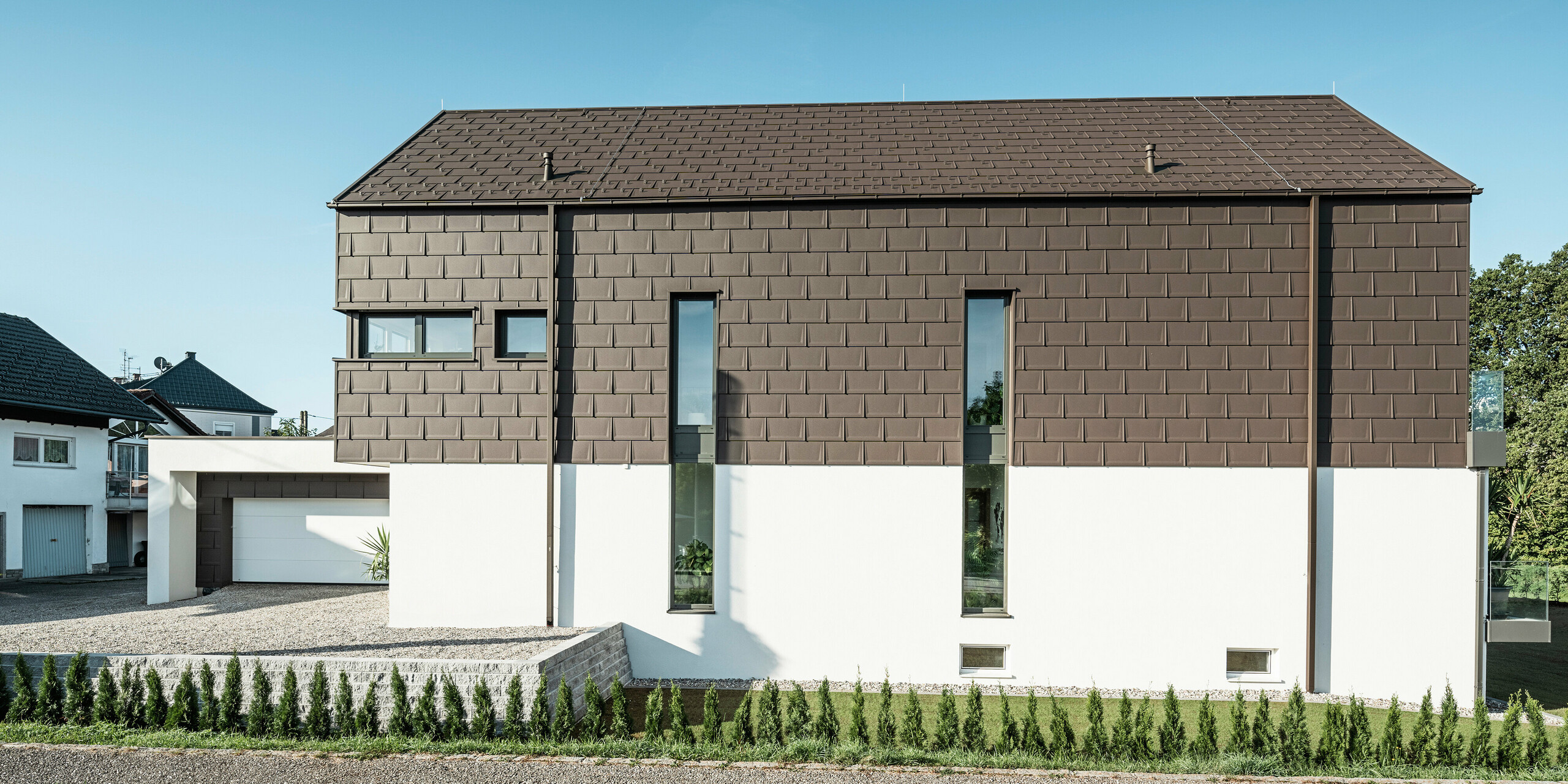 Boční pohled na rodinný dům v Neukirchenu v Rakousku se střešními panely PREFA R.16 v P.10 hnědé na střeše a na fasádě horního patra. Boční strana domu je vybavena vertikálními okny, která probíhají přes obě podlaží a zvýrazňují moderní designové pojetí domu. Jednotná struktura a jednotný barevný koncept střešních panelů R.16 nabízí výrazný vzhled. Střešní odvodňovací systém PREFA v podobě čtvercového tubusu a boxového okapu je rovněž v barvě P.10 hnědá, což umožňuje bezproblémové začlenění do současné architektury. Bílá omítková fasáda, která zdobí přízemí a garáž, dotváří harmonický celkový obraz. Mimořádnou stavbu rámují mladé cypřiše, které za pár let poskytnou soukromí kolem domu.