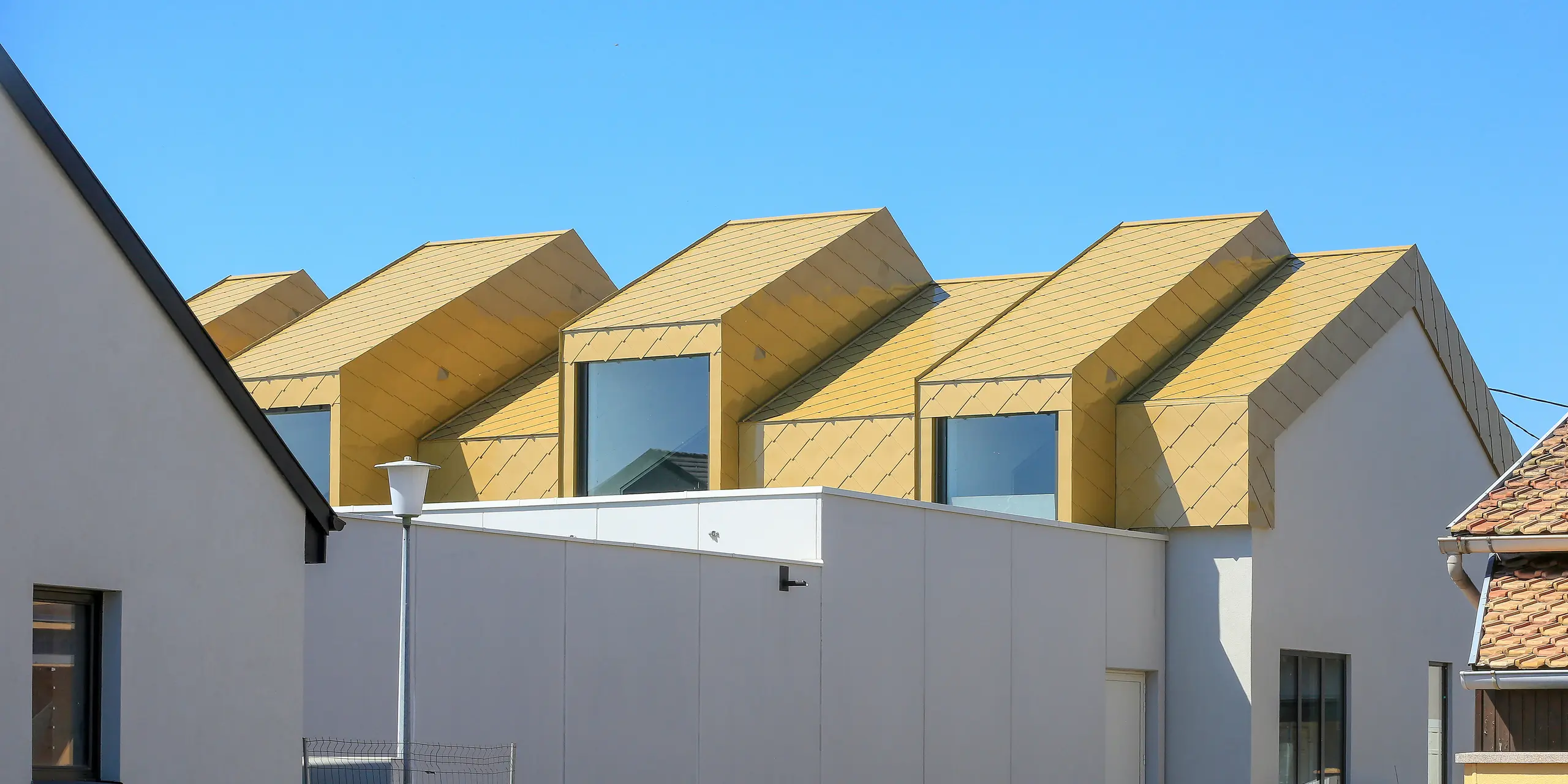 Pohled z dálky na budovu s lesklou střechou PREFA a fasádními šablonami 44 × 44 v provedení Sparkling Gold (barvě na přání)