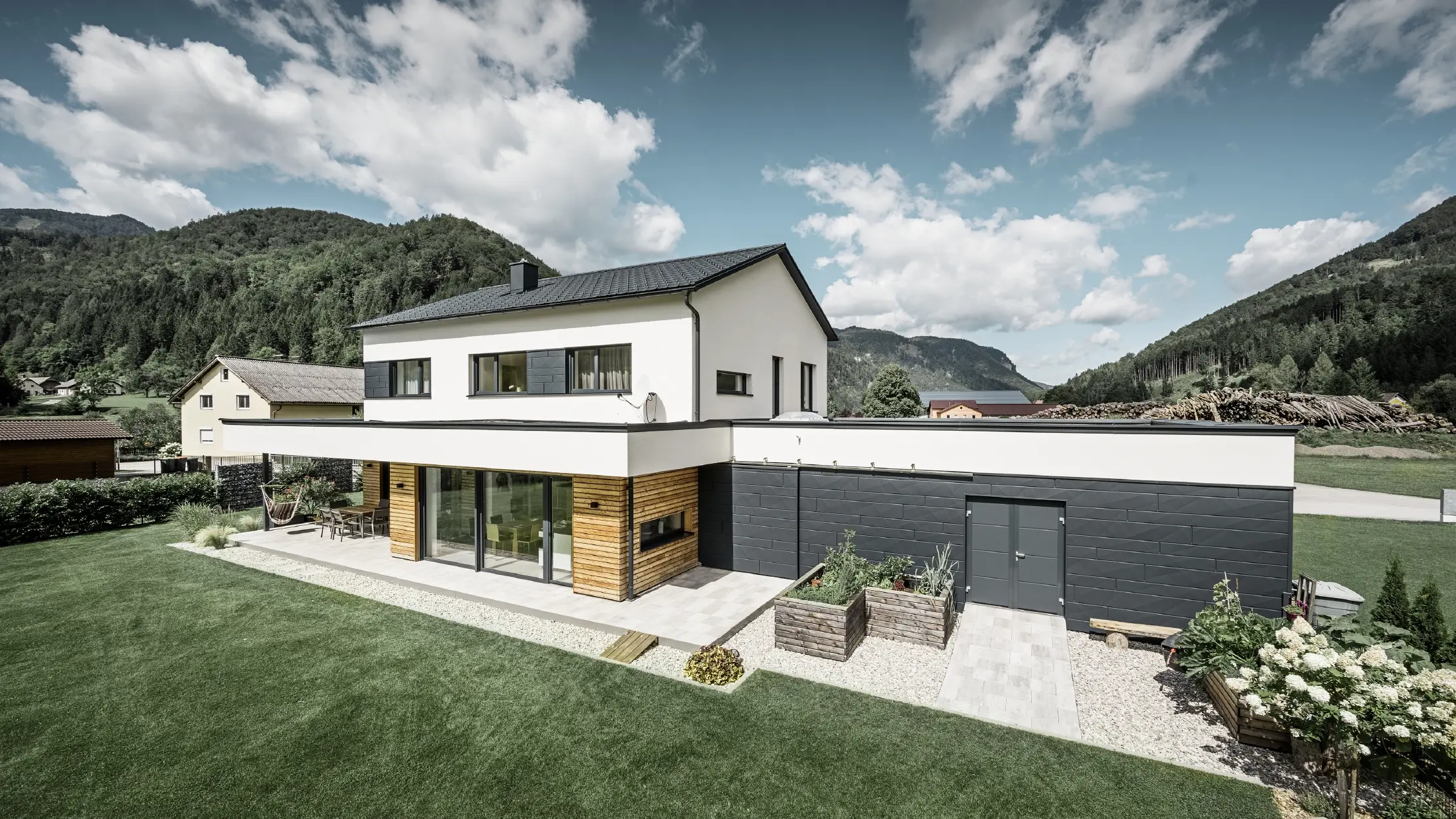  Moderní rodinný dům s bílou omítkou a PREFA Siding.X v barvě P.10 antracitová, obklopený idylickou přírodou. Tmavá fasáda kontrastuje se světlým okolím. Falcované tašky PREFA a odvodnění střechy dotvářejí odolnou konstrukci.