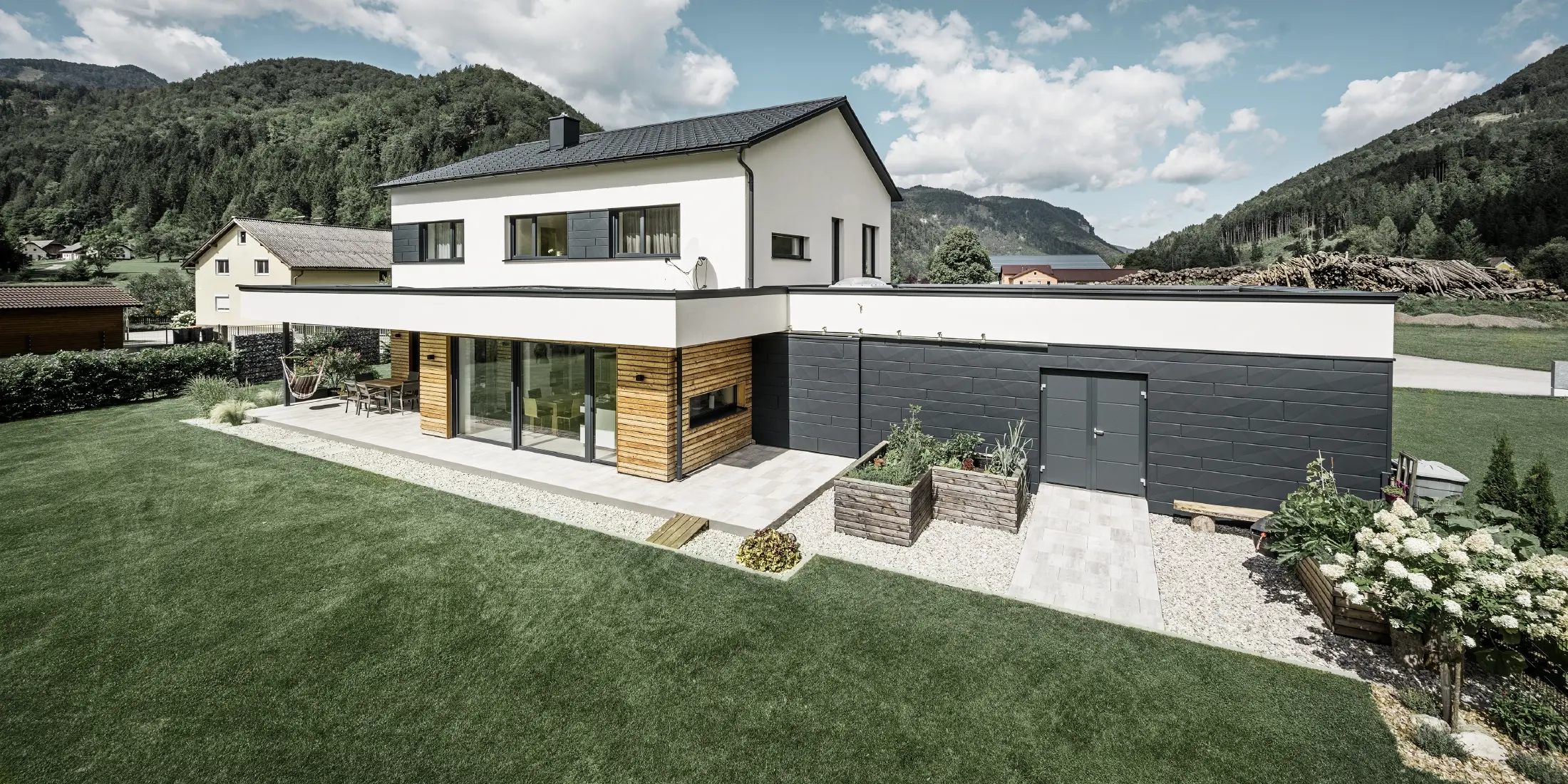  Moderní rodinný dům s bílou omítkou a PREFA Siding.X v barvě P.10 antracitová, obklopený idylickou přírodou. Tmavá fasáda kontrastuje se světlým okolím. Falcované tašky PREFA a odvodnění střechy dotvářejí odolnou konstrukci.