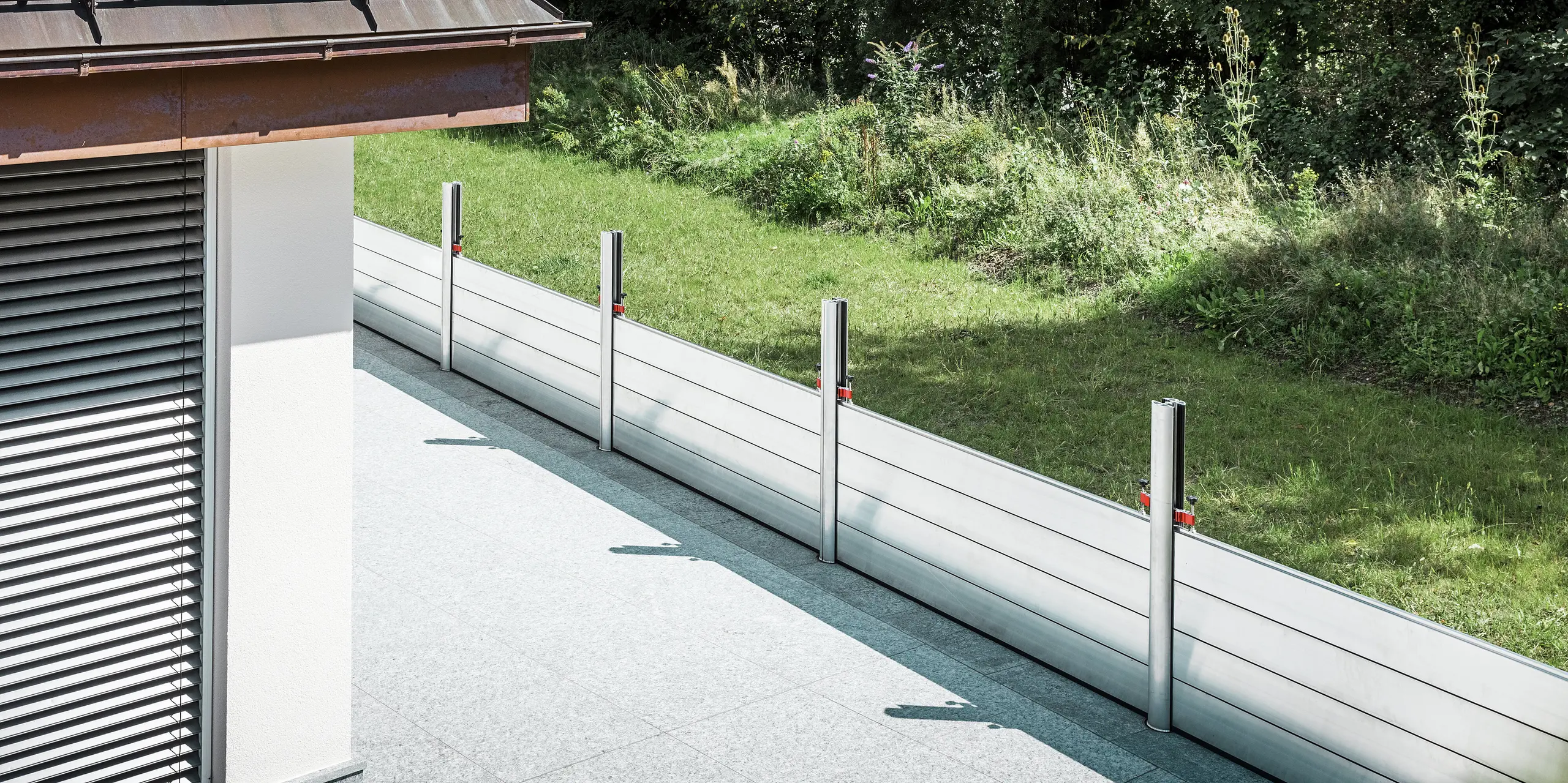 Schrägansicht des installierten PREFA Hochwasserschutzsystems entlang der Terrasse eines Wohnhauses in Tirol. Die robusten Dammbalken aus Aluminium sind zwischen fix verankerten Stützprofilen montiert, die mit Bodenhülsen fest im Untergrund verankert sind. Der mobile Hochwasserschutz bietet einen zuverlässigen Schutz vor möglichen Überflutungen aus dem angrenzenden Grünbereich. Im Vordergrund ist die gegliederte Fassade mit Jalousien zu sehen, während sich das Schutzsystem dezent und funktional in das hochwertige Wohnumfeld einfügt.