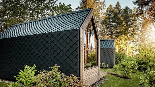 Minimalistický malý domek patřící do Secret Koi Garden u jezera Natterer se nachází v zeleni, zalitý teplým večerním světlem. Střecha a fasáda jsou kompletně obloženy drobnými střešními a stěnovými šindeli PREFA z hliníku v antracitové barvě P.10, jejichž precizní struktura připomínající rybí šupinu vytváří výrazné akcenty. Odolný a povětrnostním vlivům odolný hliník PREFA spolehlivě chrání architekturu a dokonale ladí se dřevem, sklem a okolní přírodou.