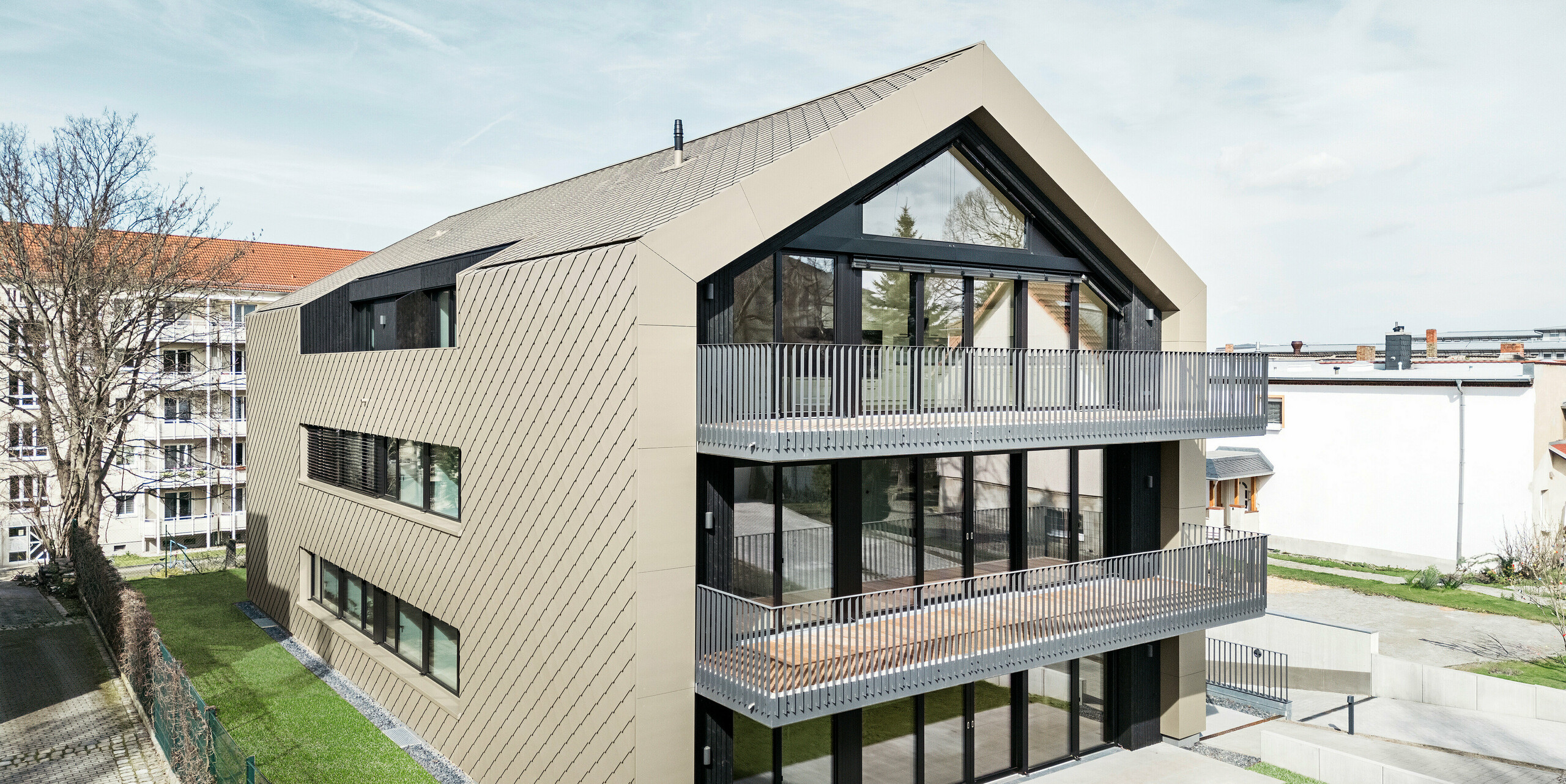 Pohled do zahrady na moderní bytový dům na Chemnitzer Straße v Drážďanech. Udržovaná zahrada je obklopena působivou architekturou. Fasáda a střecha budovy jsou opláštěny robustní falcovanou a fasádní šablonou 29 × 29 v bronzové barvě. Hliníkové šablony PREFA dodávají konstrukci elegantní vzhled. Obrázek zobrazuje prostorné balkony, velká okna a harmonické začlenění budovy do okolního prostoru.